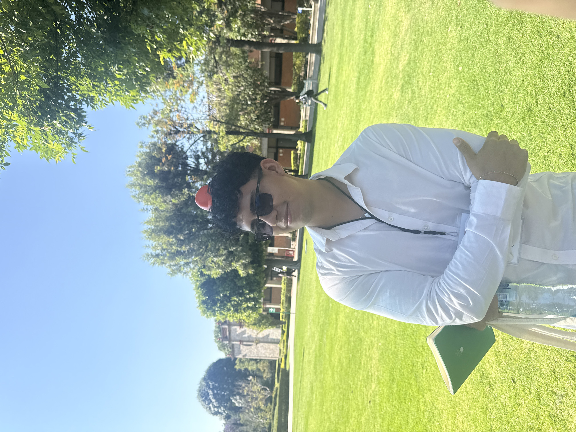 A young man stands on a lawn in San Andrés Cholula, Mexico. He wears a white shirt, sunglasses, and a red ball on his head. He is holding a green notebook and a water bottle. The background includes trees, a building, and a clear blue sky. There is green grass surrounding him.