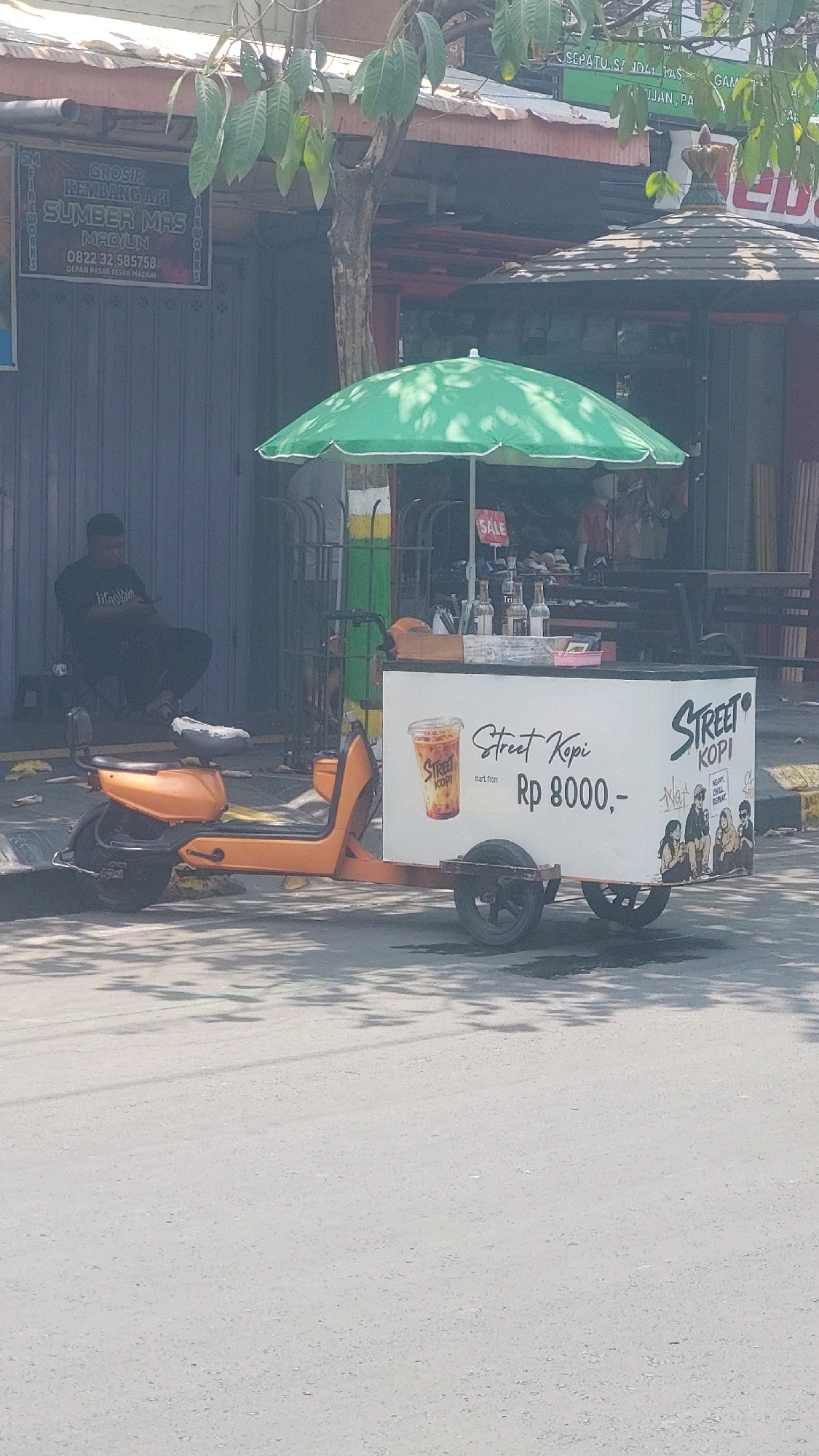 A street coffee cart, branded "Street Kopi," is parked on an asphalt road in Madiun, Indonesia. The cart is attached to an orange electric scooter. A green umbrella shades the cart. A picture of a coffee cup is on the side of the cart, along with the text "Street Kopi" and "start from Rp 8000,-". A drawing of four people sitting on a bench is also on the cart, with the text "NGAT NOOM CHILL REPEAT". In the background, a man is sitting on a stool, looking at his phone. He is wearing a black t-shirt with the words "life is pain" on it. A storefront with a rolled-down shutter is behind him. A sign on the storefront reads "GROSIR KEMBANG API SUMBER MAS MADIUN 0822 32 585758 DEPON PASAR BESAR MADIUN". The scene appears to be on a street with buildings and trees.