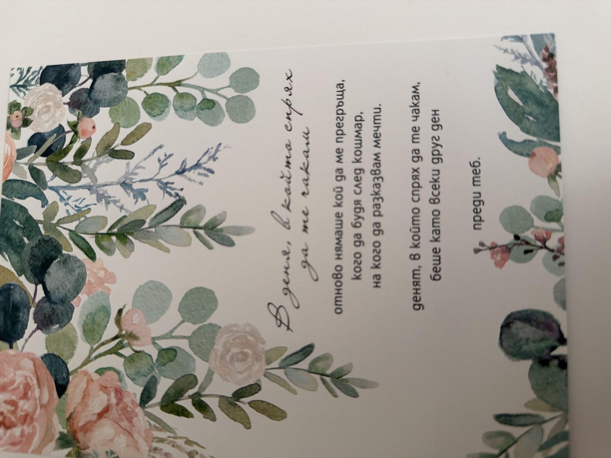 The image is a close-up, slightly angled shot of a card with a floral watercolor design. The design features pink and white roses and green eucalyptus leaves and branches, creating an elegant border at the top and bottom of the card. The text on the card is in Bulgarian and appears to be a poem or a heartfelt message. The writing is in a cursive font. The card is set against a plain, light-colored background, likely a table or surface. There is no indication of the location being Pazardzhik, Bulgaria.