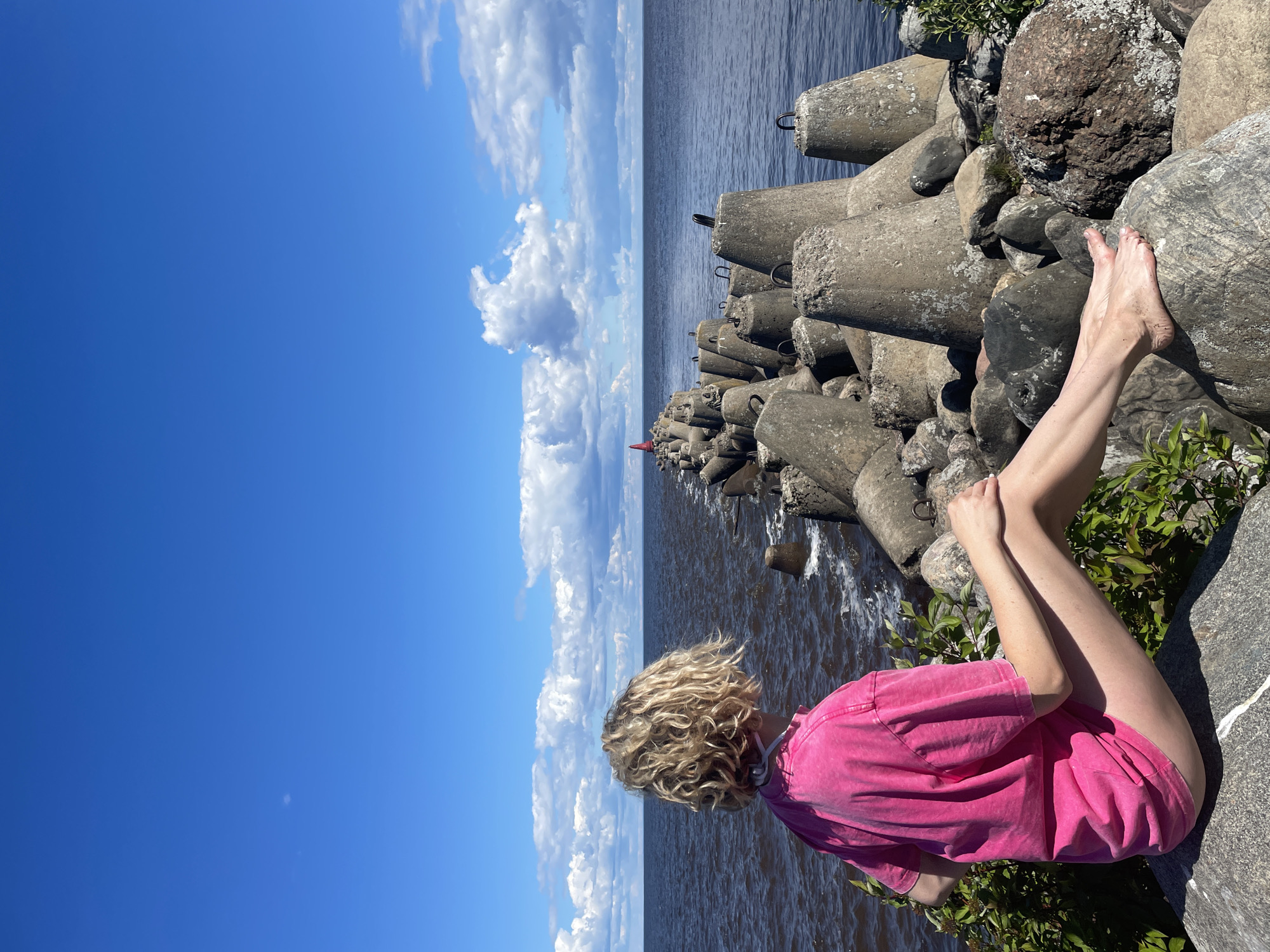 A young woman with blonde, curly hair sits on large rocks facing away from the camera, looking out at a body of water. She is wearing a bright pink t-shirt and her legs are visible. The rocks are part of a breakwater that extends into the water, made of large, concrete tetrapods. A red navigation beacon is visible in the distance at the end of the breakwater. The sky is blue with scattered white clouds. The scene is likely on the shore of Lake Beloye in Belozersk, Russia.