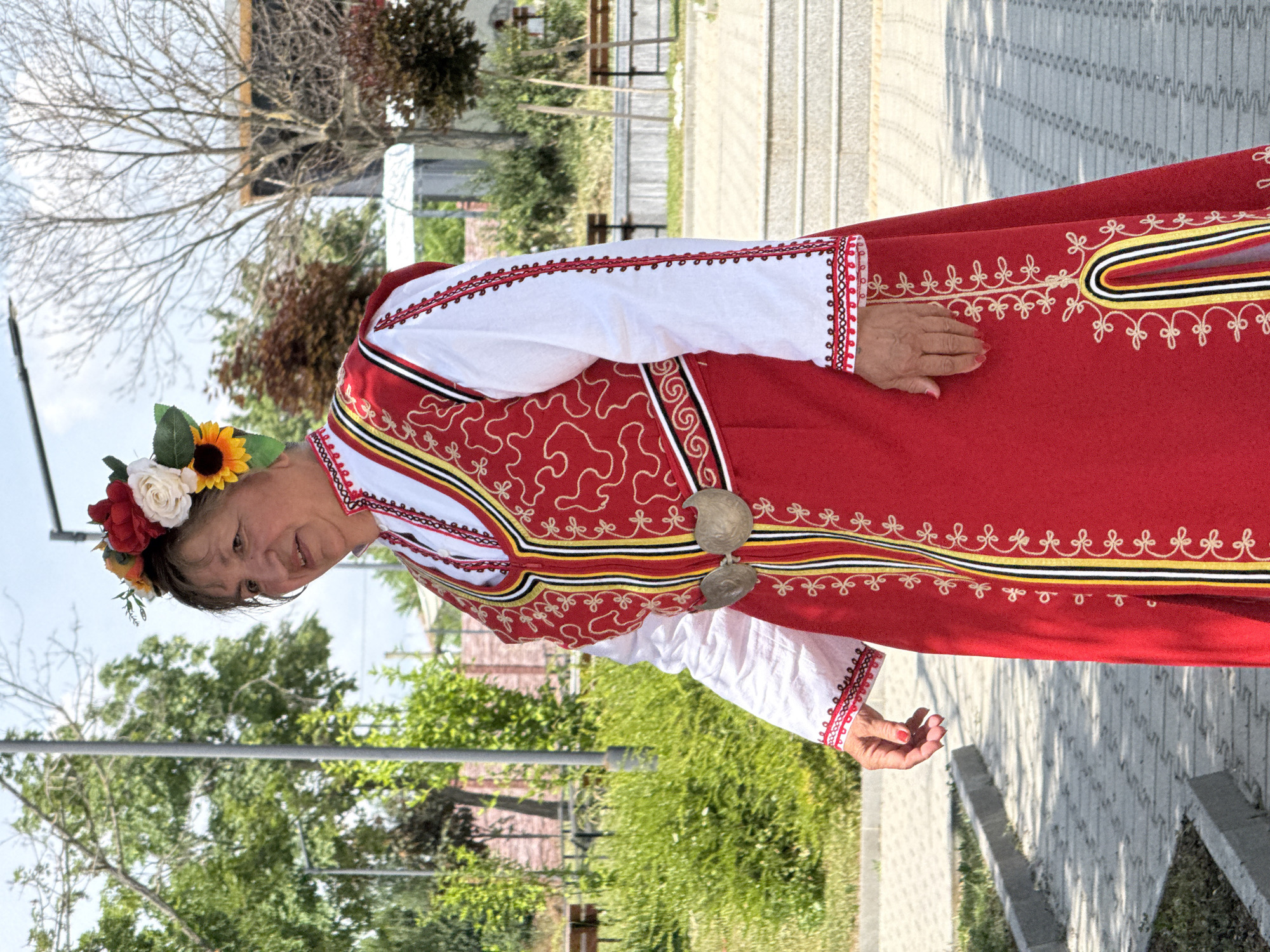 The image shows a woman in traditional Bulgarian folk costume standing outdoors. She is wearing a red outer garment with intricate embroidery and a white undergarment with embroidered sleeves. A floral headdress adorns her hair, and she has a decorative belt with silver clasps. The background suggests an outdoor public space, possibly a park or street, with greenery, trees, and paved pathways. The setting is identified as the town of Pordim, Bulgaria. The woman appears to be posing for the photograph.