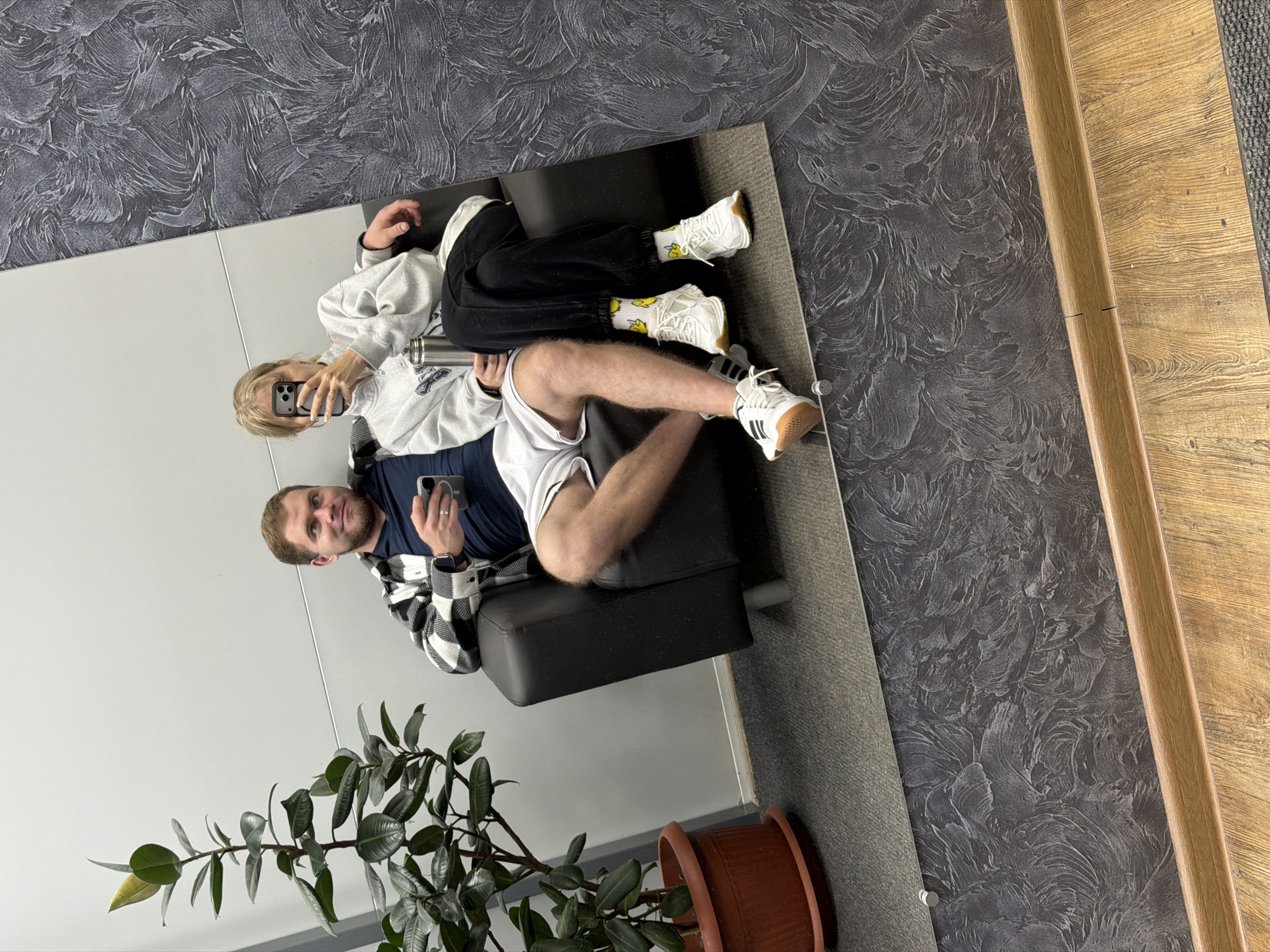 A reflection in a mirror shows a man and a woman sitting on a black chair. The man, holding a smartphone, is wearing a dark blue shirt, a black and white checkered jacket, and white shorts. The woman, also holding a smartphone, is wearing a light gray sweatshirt and black pants. Both are wearing white sneakers. The background shows a gray wall with a textured design and a wooden floor. A potted plant sits in the lower left corner of the reflection. The location is Chaikovsky, Russia.