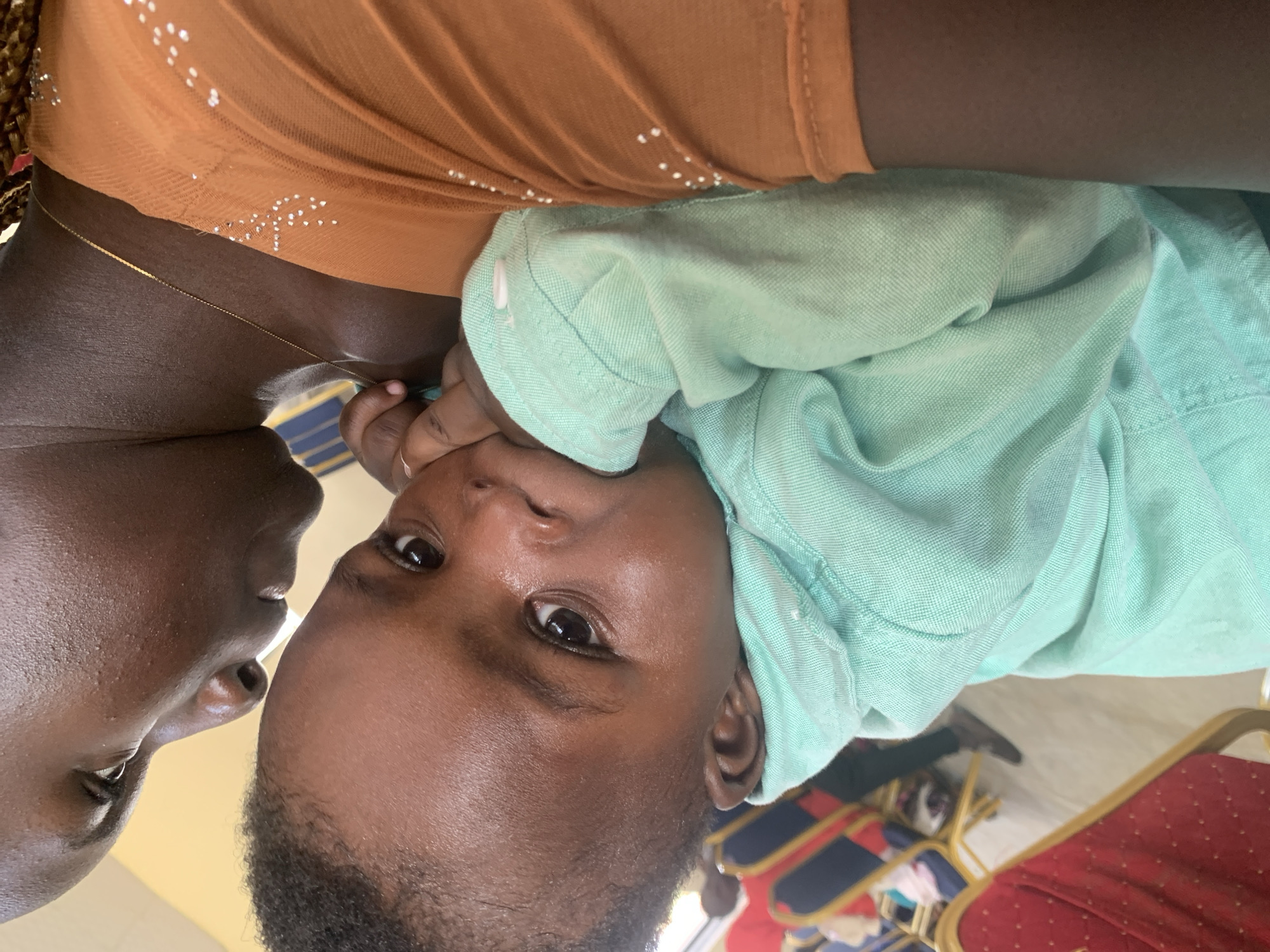A close-up, angled shot shows a Black adult holding a Black infant. The adult, wearing an orange top with rhinestones, is partially obscured by the infant. The infant, dressed in a mint green collared shirt, has its fingers in its mouth and is looking directly at the camera with wide eyes. The background is blurred, but hints of chairs with red and blue upholstery and a person in dark pants and gray shoes are visible, suggesting an indoor setting possibly a hall or event space. The lighting is warm, with the infant's face well-lit.