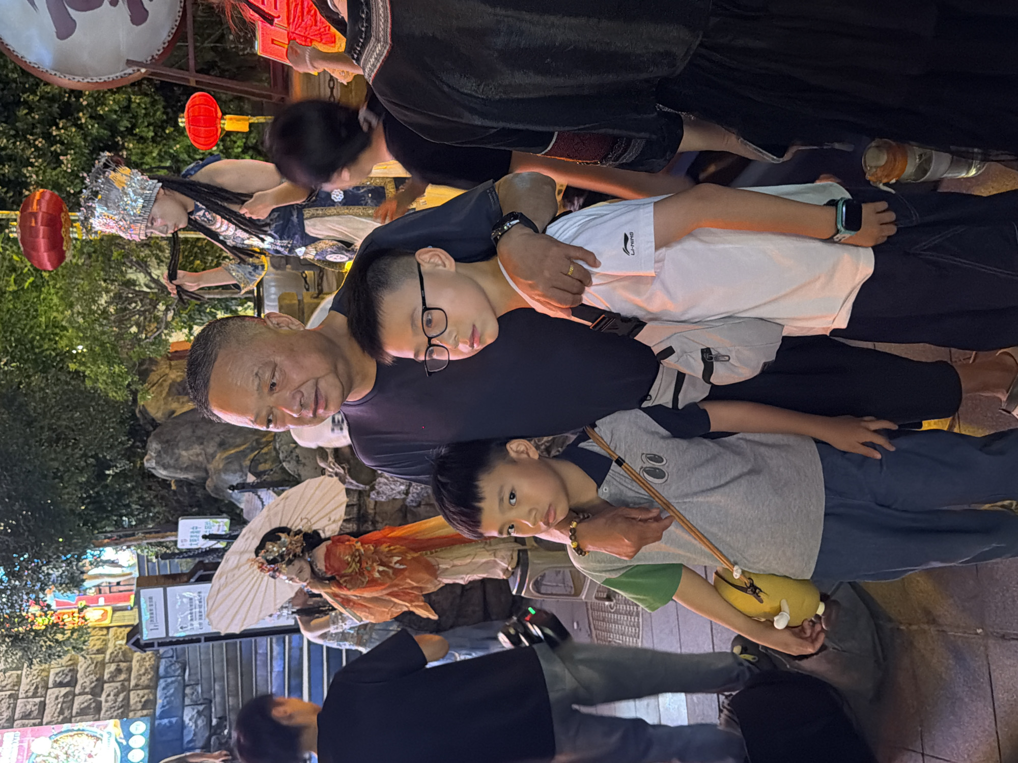 In Yuzhong District, China, at night, a group of people stand near a street performance. In the foreground, an adult male wearing a dark t-shirt is partially visible on the right, with his arm around the shoulder of a young boy. The boy, wearing a gray and green t-shirt and blue pants, holds a yellow plush toy. To the left of the boy is another young boy wearing a white t-shirt and dark pants, with glasses. Behind them, several other people are visible, including an adult female dressed in traditional costume with an elaborate headdress and dark braids, holding what appears to be a prop. A large red lantern hangs overhead, and the background suggests a bustling street or market area with stone structures and foliage.