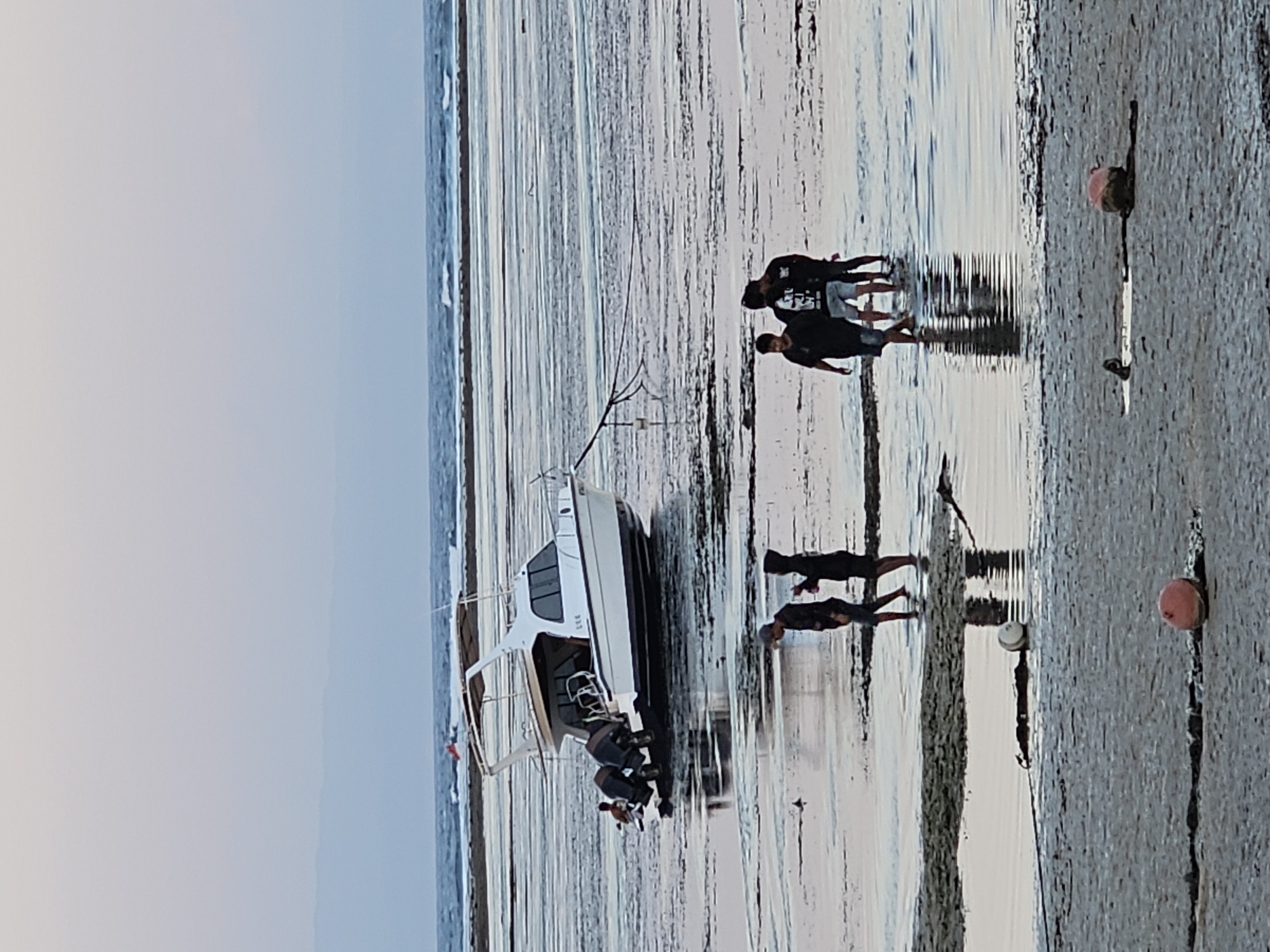 A wide shot captures a coastal scene at what appears to be low tide in Denpasar, Indonesia. A white motorboat, with two outboard motors, rests on the shallow seabed. Several people are visible in the water and on the exposed seabed around the boat and further in the distance. In the foreground, two buoys, one red and one white and partially submerged, lie on the wet sand. The sky is overcast with a pale gradient from light grey to a soft pink hue. The sea in the background has small waves.