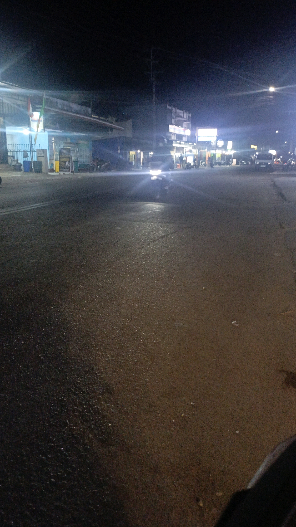 The image depicts a street scene at night in Taliwang, Indonesia. A motorcycle is prominent in the foreground, its headlights casting a bright glare, suggesting it is either moving or stationary. The road surface appears wet and reflective, indicating recent rain or damp conditions. In the background, buildings with illuminated signs line the street, suggesting a commercial or populated area. Several other vehicles, possibly motorcycles and a van, are visible further down the road. People are present on the sidewalks and near the buildings, though their actions are not clearly discernible due to the low light and distance. The overall atmosphere is dark, with artificial lights from buildings and vehicles creating illuminated pockets within the scene.