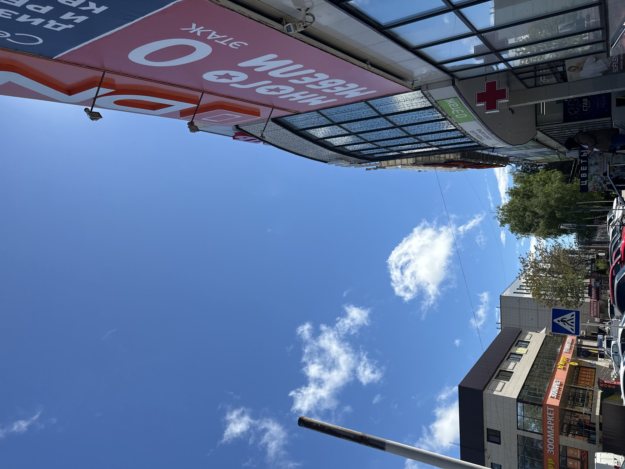 The image shows a street scene in Stavropol, Russia. Visible are commercial buildings, including one with a large red sign displaying "МНОГО МЕБЕЛИ 0 ЭТАЖ" and another marked "300МАРКЕТ" with advertisements for various goods. A pharmacy with a red cross logo is also present. There are trees along the street. Cars are parked alongside the road. The sky is bright blue with scattered clouds. Street signs and other smaller details such as awnings and streetlights are visible.