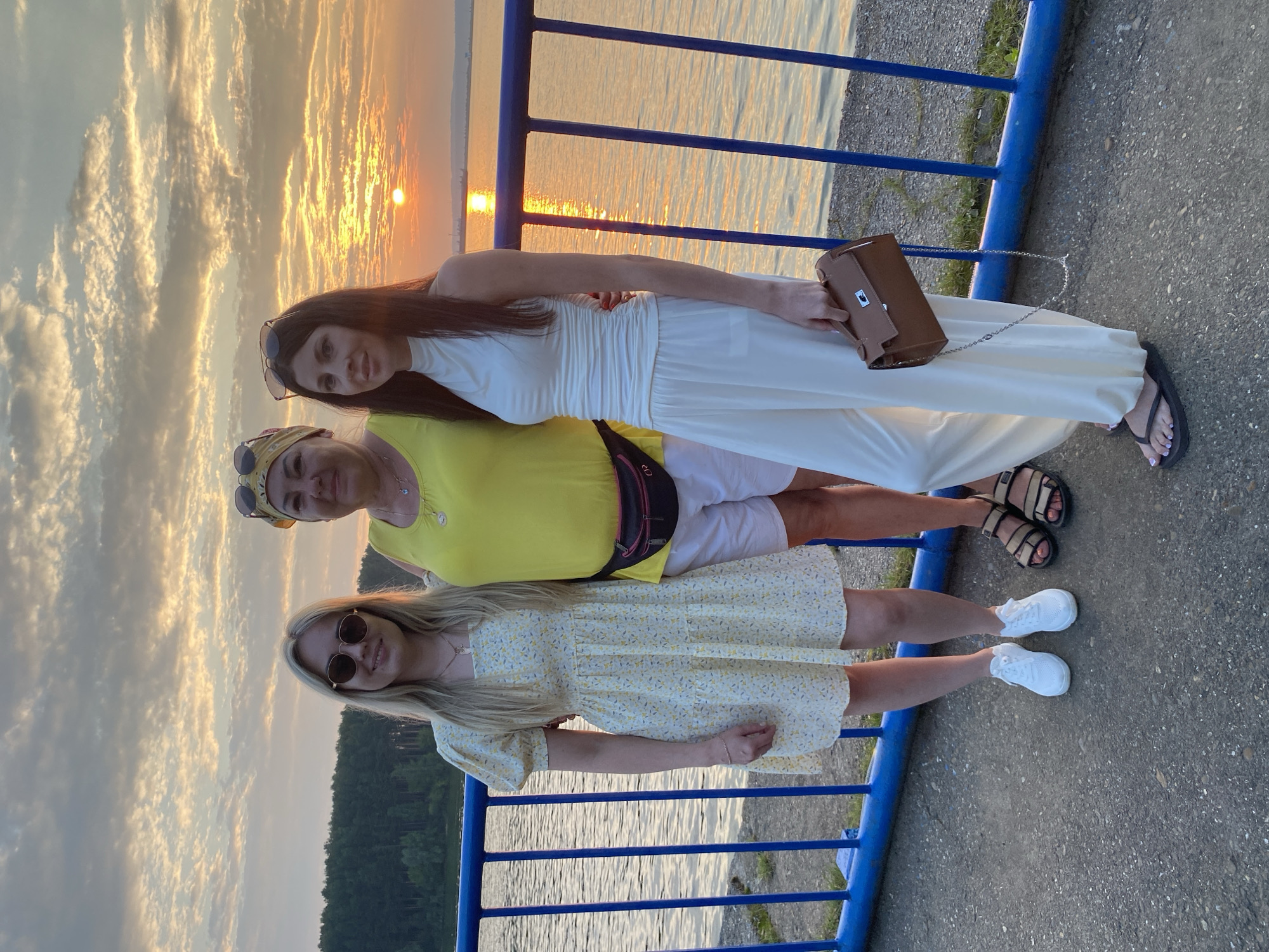 Three individuals, two women and a mature woman, stand together on a paved walkway next to a blue metal railing. The background features a body of water with a distant shoreline of trees, and a sunset with dramatic cloud formations and the sun low on the horizon, casting a warm glow on the scene. The mature woman in the center wears a yellow sleeveless top and white shorts, accessorized with a yellow bandana and a fanny pack. To her left, a young woman in a yellow floral print dress and white sneakers stands with her left hand on the railing. To the mature woman's right, a young woman in a white sleeveless top and white wide-leg pants holds a brown handbag. The setting appears to be a waterfront promenade or embankment, possibly within the city of Chaikovsky, Russia, given the presence of a large body of water and forested areas.