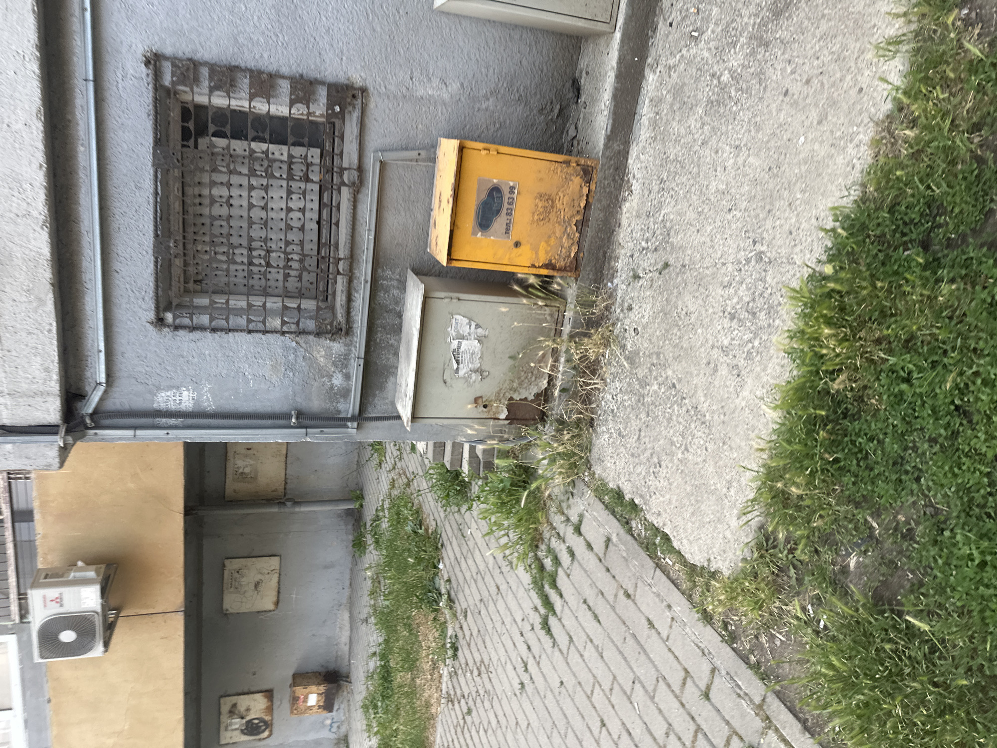 This photograph captures a quiet, weathered urban scene, likely an alleyway or a side passage between buildings. No people are visible.
The setting consists of several building walls, a paved walkway, and patches of overgrown grass. The primary wall on the left is grey, with a rough, textured finish. Embedded in this wall is a rectangular window opening, protected by a heavily rusted, ornate metal security grate. Below and to the right of the window, a bright yellow electrical or utility box is mounted on the wall. It shows significant signs of rust and wear, with peeling paint. A dark, possibly stylized logo is visible on its front, along with white text that reads "Тen.: 63 63 99", likely indicating a phone number. Below the yellow box is another, lighter-colored utility box, also damaged and displaying remnants of an illegible white sticker.
To the right of the boxes and wall, a grey paved walkway, made of rectangular pavers, extends into the distance. Alongside the pavement, a strip of concrete and dirt is heavily overgrown with green grass and weeds, suggesting a somewhat neglected area. Further into the scene, a section of a light yellow/beige building wall is visible, featuring a white Mitsubishi outdoor air conditioning unit. Another grey wall in the background has several square panels or covers mounted on it. Various pipes and conduits run along the walls.
The scene depicts a clear, bright daytime, with no indication of specific weather conditions other than dry. The overall impression is one of aging infrastructure and ordinary urban details, typical of an older residential or industrial area in Burgas, Bulgaria.