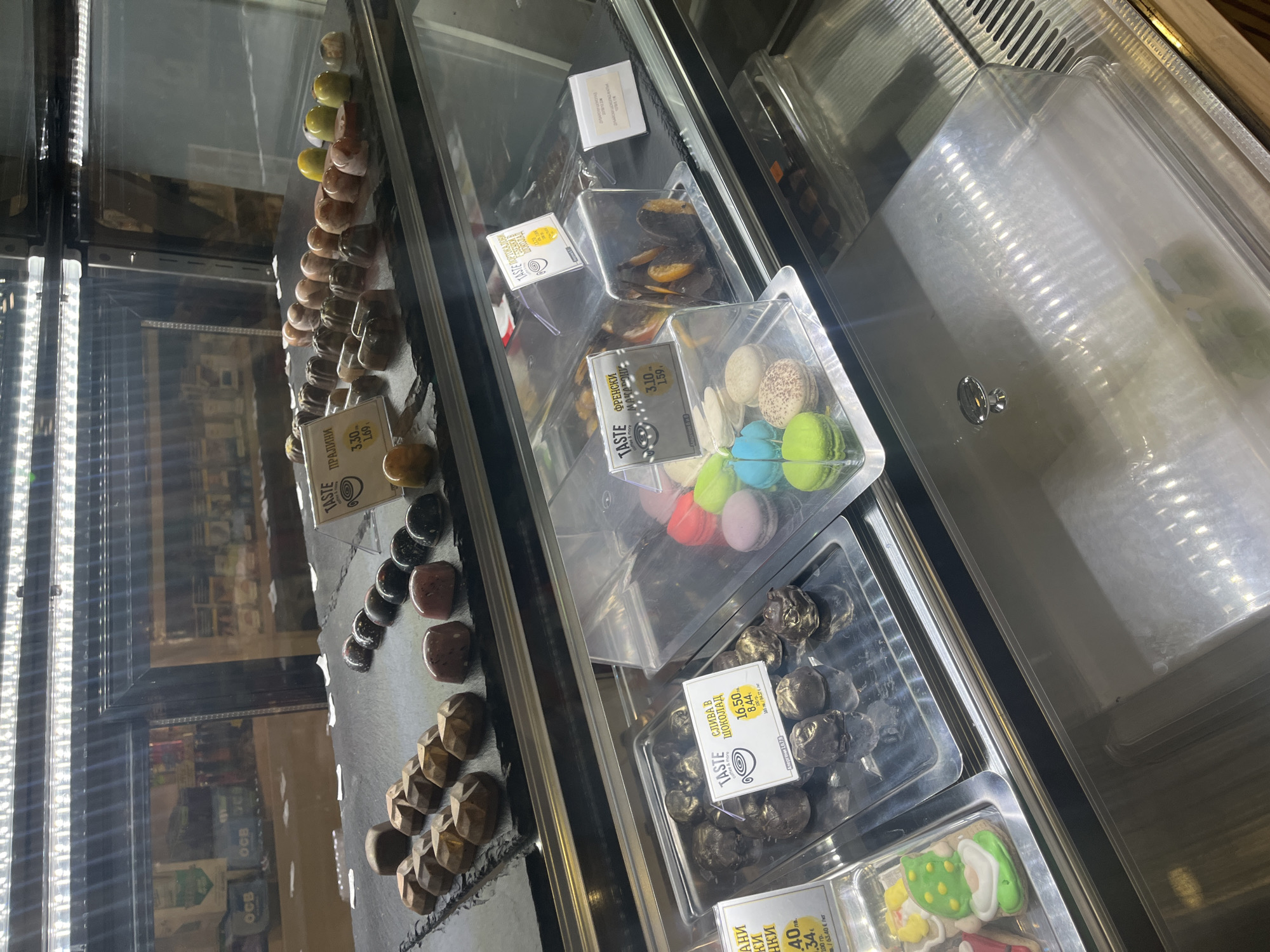 The image shows a display case filled with a variety of confectionery items, possibly in a shop in Plovdiv, Bulgaria.
The leftmost section contains rows of individual chocolates of different shapes and colors, placed directly on a dark surface. A small price tag is visible, labeled "TASTE ПРАЛИНИ" with prices of 3.30 and 1.69.
The middle section holds plastic containers of desserts. One container has colorful macarons, another contains fruits or plums covered in chocolate, with a "TASTE СЛИВА В ШОКОЛАД" price tag showing 16.50 and 8.44. A container with dried apricots is also visible.
The rightmost section shows more plastic containers, one filled with small round chocolates, labeled "TASTE ОРАНЖЕВИ". The bottom right corner contains biscuits decorated with green frosting.
The background is blurred, showing reflections on the glass of the display case.