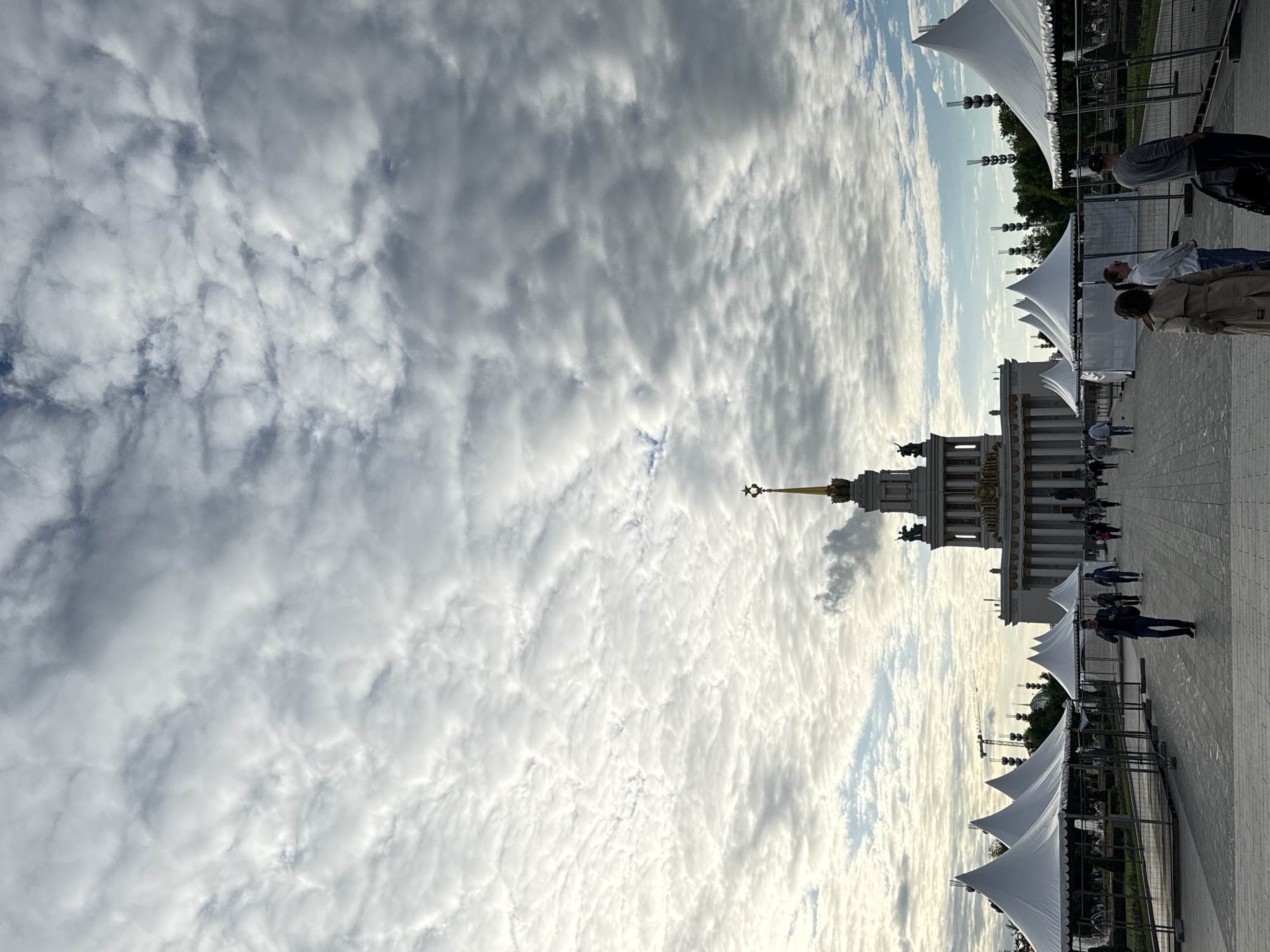 This outdoor image, taken in Moscow, Russia, captures a grand architectural setting under a dramatic sky, likely at the Exhibition of Achievements of National Economy (VDNKh). The most prominent feature is a magnificent classical building on the right side of the frame, featuring columns, arches, and a tall central tower culminating in a golden spire topped with a star, characteristic of the Main Pavilion at VDNKh.
In the foreground and extending towards the midground on the right, several white, pointed tents or canopies are set up along a wide, paved promenade. Metal fences delineate pathways or event areas around these tents. Behind the tents, some decorative lighting fixtures on poles are visible, and in the distance, outlines of structures, possibly construction cranes, can be faintly seen.
Numerous people are scattered across the paved area, casually walking or standing near the tents and the main building. They appear to be visitors or attendees, dressed in everyday attire, suggesting a public event or a busy day at the complex.
The time of day appears to be late afternoon or early evening, indicated by the quality of light which casts some soft shadows and illuminates the clouds. The sky dominates a large portion of the image, filled with an expansive display of textured, broken clouds, likely altocumulus or stratocumulus, creating a dynamic and visually interesting backdrop. Despite the cloud cover, there are patches of clear sky, allowing sunlight to break through, suggesting fair weather. No discernible text is visible in the image. The scene conveys a sense of public activity and urban grandeur.