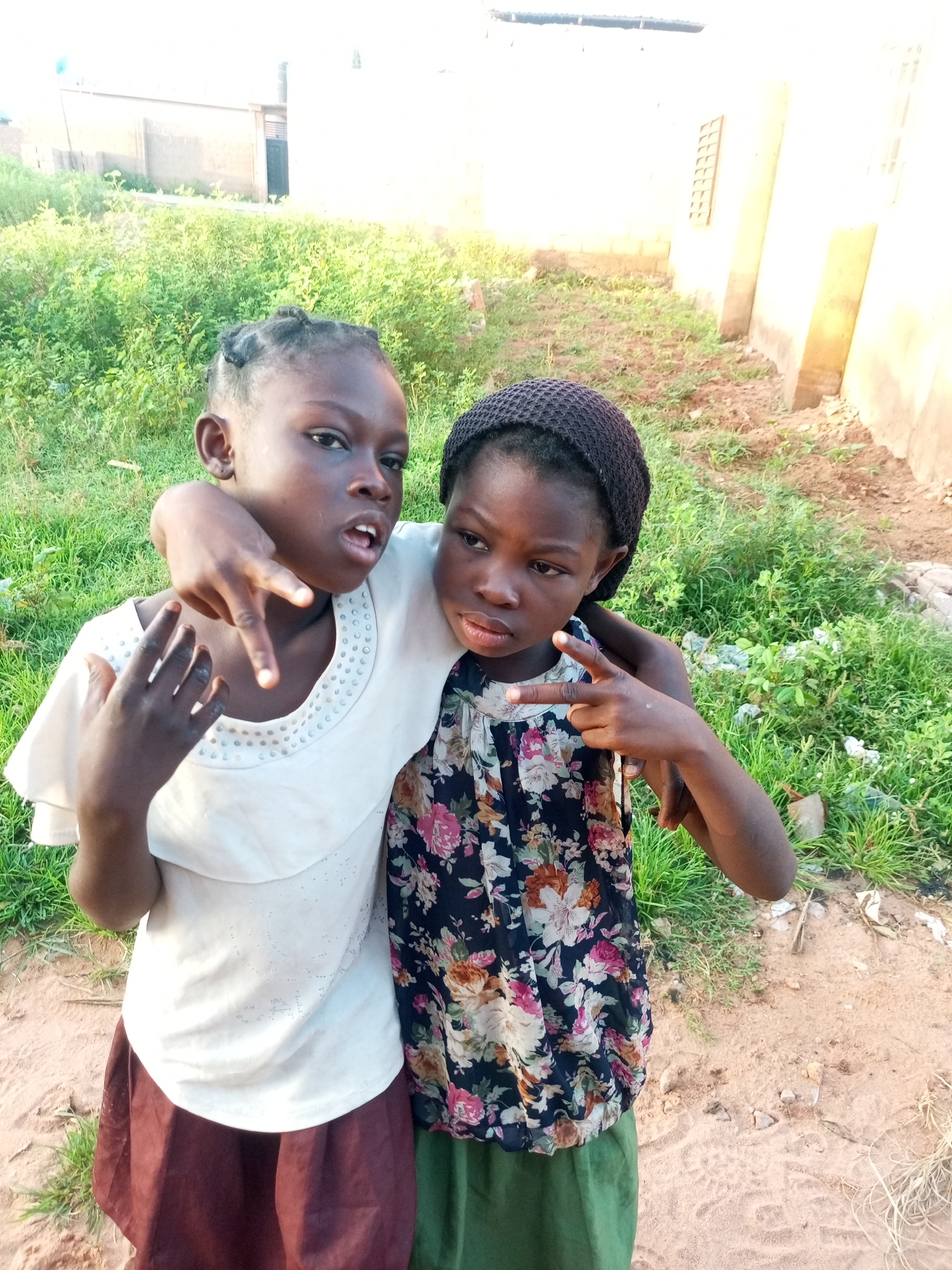 Two young girls are posed closely together outdoors, with one girl's arm draped around the other's shoulders. The girl on the left, wearing a white shirt and a brown skirt, has her left hand extended with her index and middle fingers pointing upwards in a gesture. She has braided hair. The girl on the right wears a dark floral-patterned top and a green skirt, and has a dark head covering. Both girls appear to be looking towards the camera. They are standing on sandy ground with green vegetation and buildings visible in the background. The environment suggests a somewhat undeveloped area in the city of Tempure, Nigeria, likely a residential or semi-rural setting. The lighting suggests it is daytime.