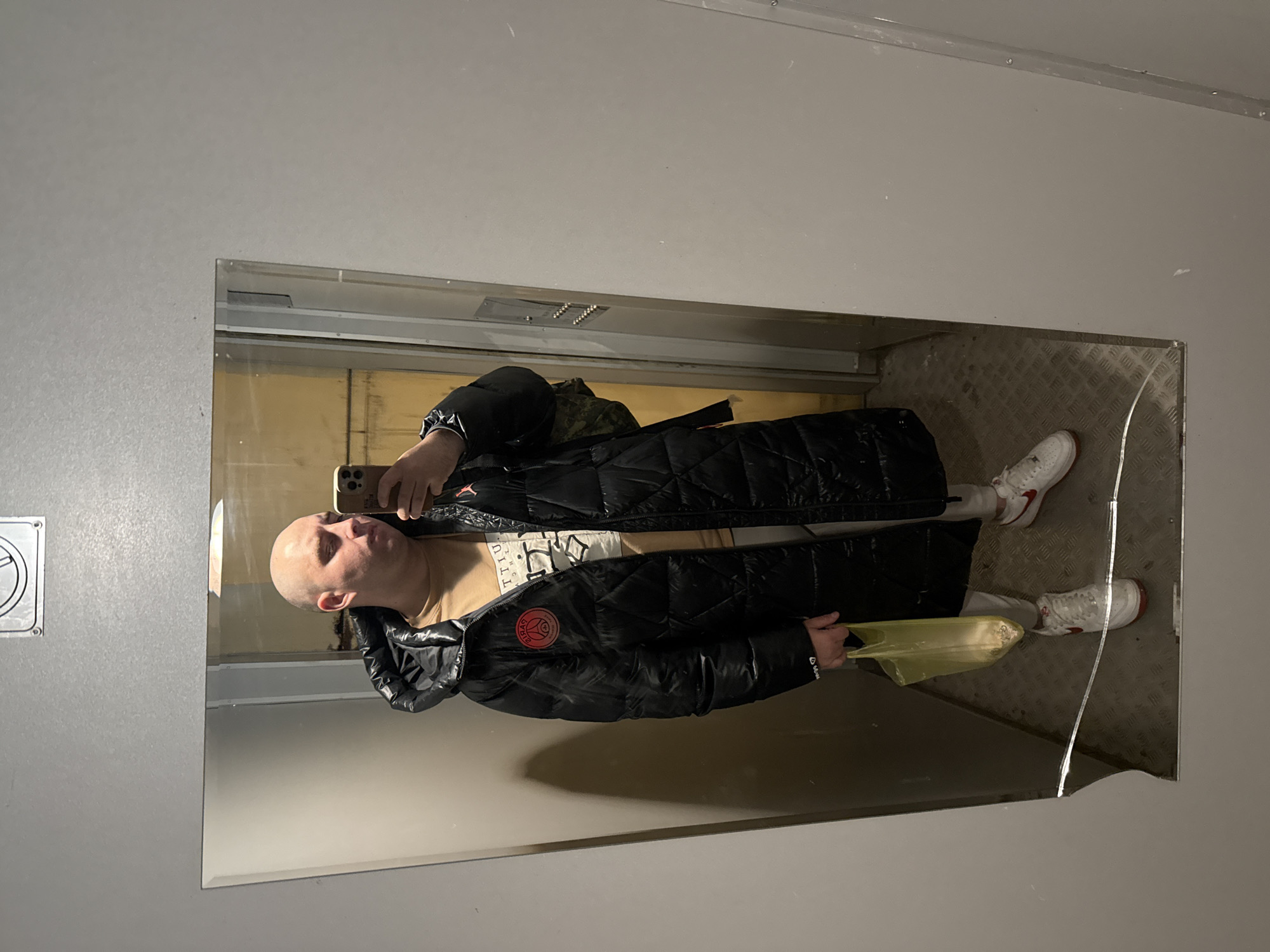 A bald man is taking a mirror selfie in what appears to be an elevator. He is wearing a black, long, puffy jacket with a logo on the left chest, a beige t-shirt with printing, light-colored pants, and white sneakers with red accents. He is holding a yellow object in his left hand. The mirror is cracked in the bottom right corner. The elevator interior is grey. There is a no-smoking sign visible in the upper left corner of the mirror. The location is Pleven, Bulgaria.