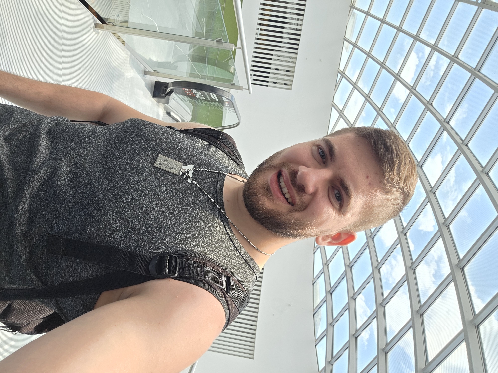 A man is standing inside a modern building with a large, grid-patterned glass ceiling revealing a view of the sky with clouds. He has fair skin, short blonde hair, a beard, and is wearing a gray patterned tank top with a black backpack. He is looking at the camera and smiling slightly. In the background, there are white walls and horizontal stripes of light, and part of a green and silver elevator or platform can be seen. The location is likely in the city of Plovdiv, Bulgaria.