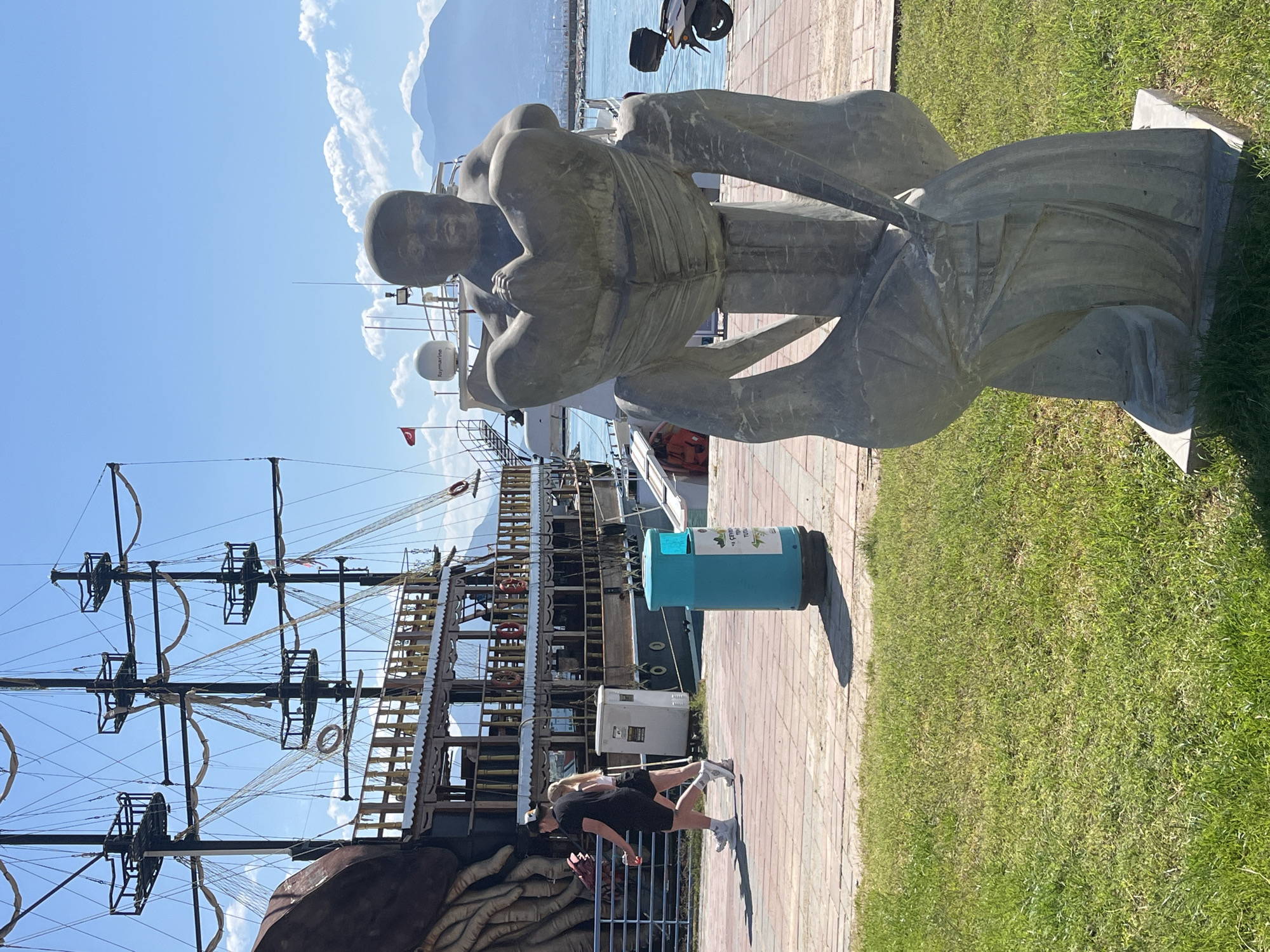 A statue of a seated man is in the foreground, on a brick walkway with grass to the right of it. In the background is a ship with a mast and rigging, other boats and ships, and a mountain range on the horizon. People are walking on the path. A trash can is next to the walkway. The sky is clear and blue. This is Alanya, Turkey.