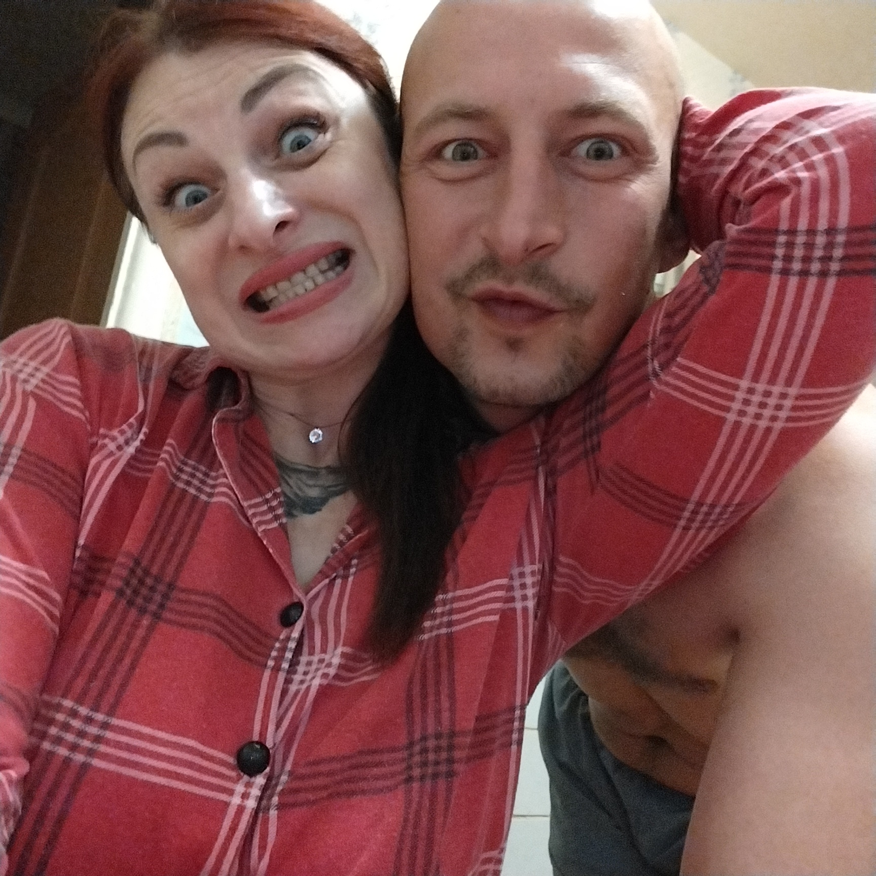A medium close-up selfie captures two individuals, a woman and a bald man, posing playfully. The woman, positioned on the left, has reddish-brown hair and is making a funny face with her eyes wide open and teeth gritted. She is wearing a red and white plaid shirt. The man, on the right, has a bald head, a mustache, and is also making a surprised expression with his eyes wide. He is leaning his head against the woman's head and his arm is wrapped around her, wearing a red and white plaid shirt as well. The background is indistinct, showing a wall and possibly a door frame. The lighting appears to be indoor, creating some highlights on their faces and shoulders.
