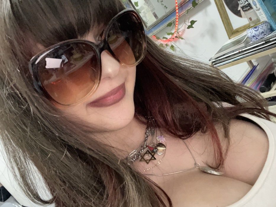 A woman with brown hair and bangs wears oversized sunglasses with an ombre tint. She has light skin and wears a dark lipstick. A layered necklace with charms, including a Star of David, drapes across her chest. She wears a white top. In the background, there are shelves with various items, including a framed picture, books, and decorative objects, with a plant vine hanging down.