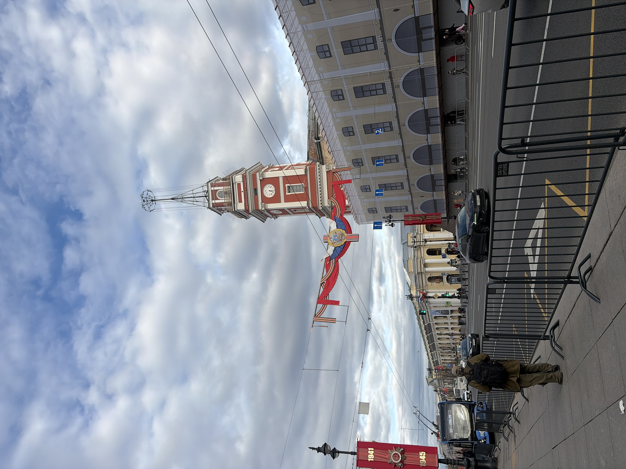 Here is a detailed description of the image:
**Location:** The image is taken in Saint Petersburg, Russia. The tall, red and white tower with a clock is the tower of the Naval Headquarters. The yellow building to the right of the tower is likely part of the Winter Palace complex or another building along Palace Square.
**Environment:** The weather appears overcast, with a sky filled with gray and white clouds. The scene is urban, featuring buildings, streets, and pedestrian walkways.
**Objects:**
* A tall red and white tower with a clock is prominent in the image.
* Red banners are strung across the street, likely for a holiday or celebration. They feature decorations and dates.
* A metal fence separates the pedestrian area from the street.
* Several cars are visible on the street.
* A street lamp is visible in the foreground.
**People:**
* A person is walking on the sidewalk, wearing a green jacket and backpack. The person is walking away from the camera.
**Actions:**
* People are walking on the sidewalk in the background.
* Cars are driving on the street.
