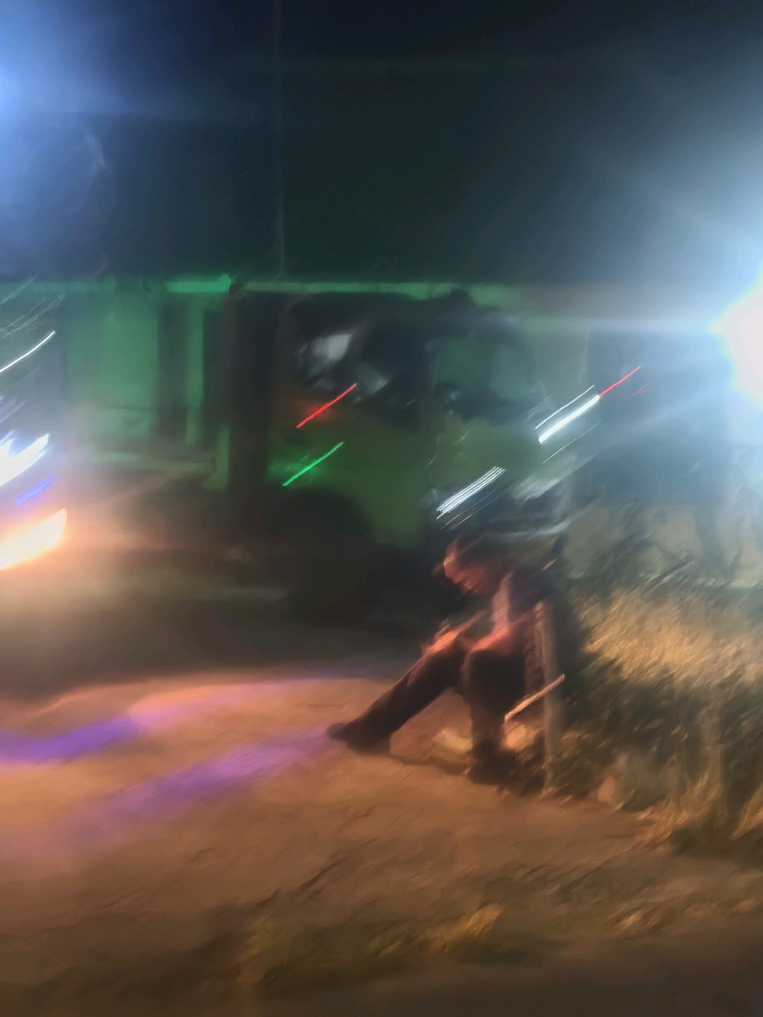 In Kroya, Indonesia, at night, a blurry scene unfolds on a dirt road. A person, appearing to be male, sits slumped against a post, leaning their head on their arm. They are wearing dark clothing. Behind them, a green vehicle, possibly a truck, is partially visible, with streaks of red and green light emanating from it. The ground is illuminated by various colored lights, including pink, purple, and orange, casting long, distorted shadows. The overall atmosphere is hazy and appears to be captured with a moving camera.