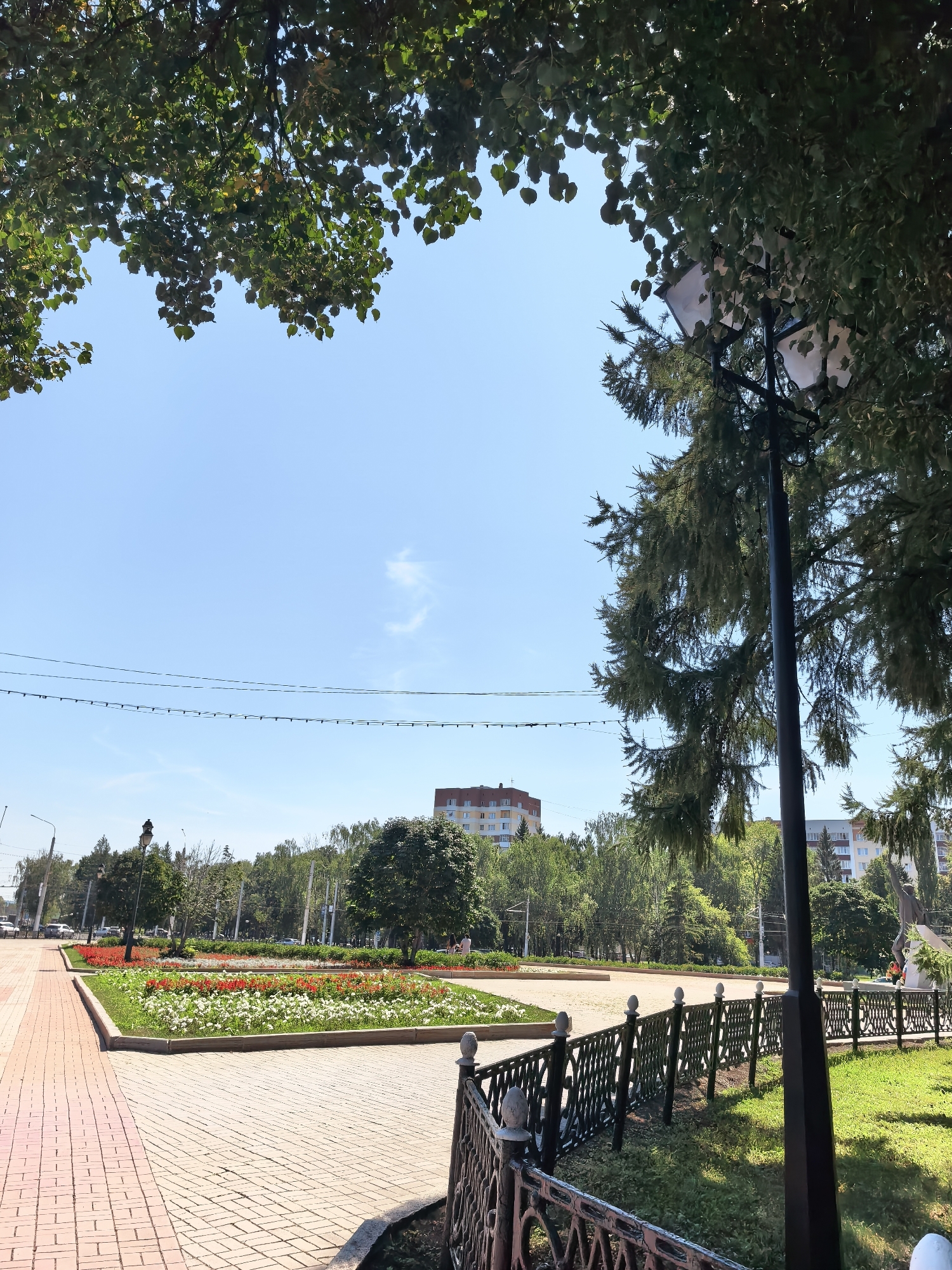 The image shows a sunny day in a city park. In the foreground, a brick pathway leads to a manicured flowerbed filled with red and white flowers. A black metal fence runs along the edge of the park, separating the pathway from a grassy area. Trees, including tall evergreen trees, line the park, casting shadows on the ground. In the background, apartment buildings and other city structures are visible amidst lush greenery. The sky is clear blue with a few wispy clouds.
This image appears to be taken in Sterlitamak, Russia, likely within a public park area given the presence of pathways, flowerbeds, benches, and greenery. The overall scene suggests a well-maintained urban green space on a pleasant day. There are no people clearly visible or engaged in any significant action within the frame. The focus is on the landscape and urban elements.