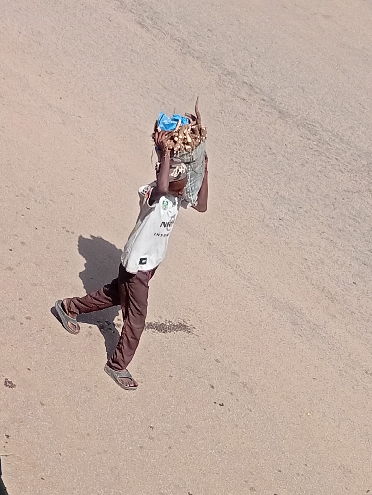 An individual, appearing to be a young male, walks across a sandy, dusty surface, likely a road in Bukuru, Nigeria. He carries a large bundle of goods on his head, wrapped in a patterned cloth and a blue plastic bag. He is wearing a white sleeveless t-shirt with writing and a logo, and dark brown trousers. His footwear consists of dark-colored sandals. The sun casts a distinct shadow to his left. The overall scene depicts a common sight of commerce or transport in an outdoor, possibly arid, urban environment.