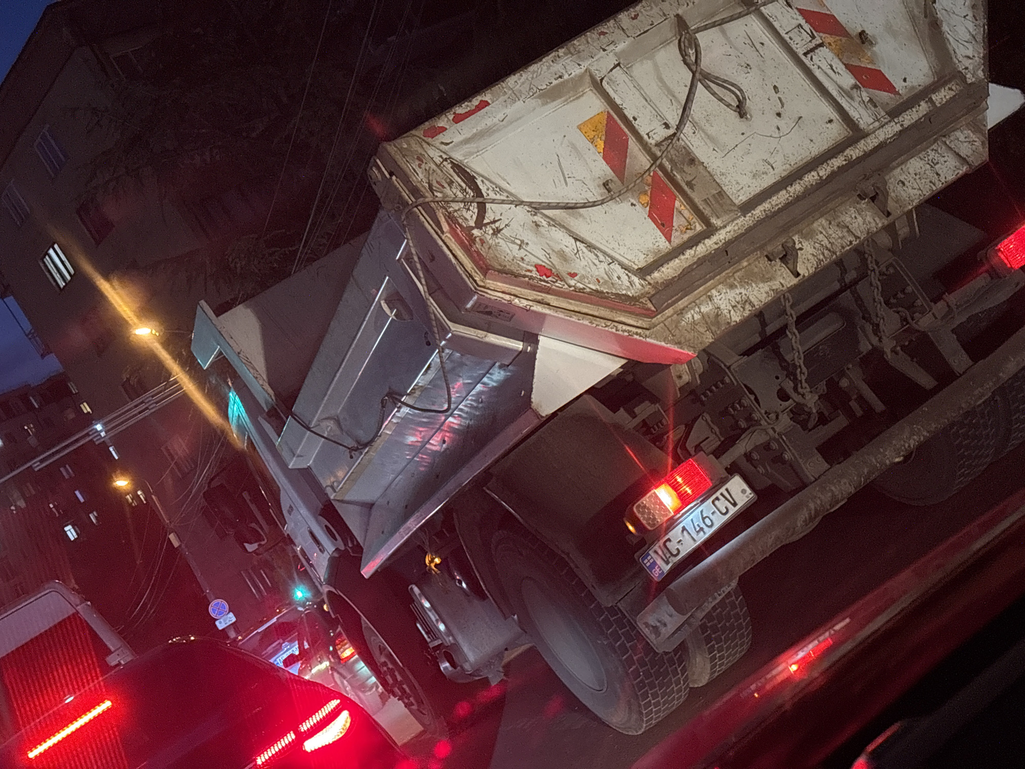 The image captures an urban street scene at night, viewed from inside a vehicle. The primary subject is a large, light-colored dump truck, seen from its rear-right side. The truck's bed appears well-used and dirty, with visible chains, ropes, and reflective hazard markings in red, white, and yellow. Its rear features illuminated red taillights and a license plate that reads "GE" with the Georgian flag icon, followed by "VC-146-CV".
In the foreground to the left, the bright red taillights of another vehicle are prominent, indicating either stopped or slow-moving traffic. In the background, dark multi-story buildings with some illuminated windows are visible. Streetlights cast bright yellow beams, and a green traffic light is visible further down the street. A blue circular street sign with a white diagonal bar is also present. The scene is illuminated by various artificial light sources against a dark night sky. No people are discernible. The visible license plate confirms the location as Tbilisi, Georgia.
