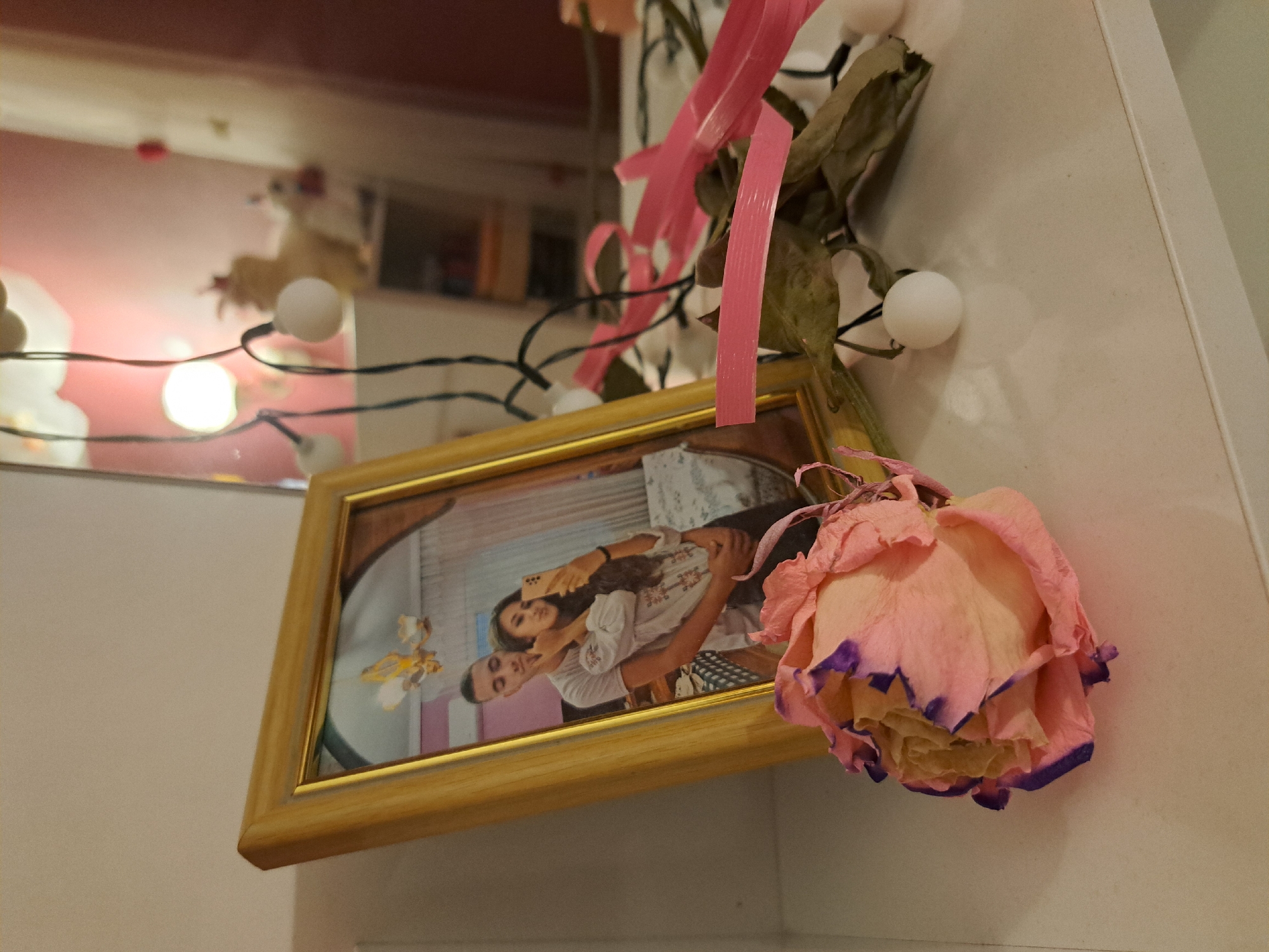 A framed photograph of a young couple is displayed on a white surface. The couple is posing for a selfie, with the man embracing the woman from behind. In the foreground, a dried pink rose with purple accents lies on the surface. Decorative white lights are strung behind the photograph and rose. The background is blurred, but appears to be a room with pink walls and a mirror reflecting a chandelier and a decorative figure. The overall scene is intimate and personal, suggesting a domestic setting in an unknown location.