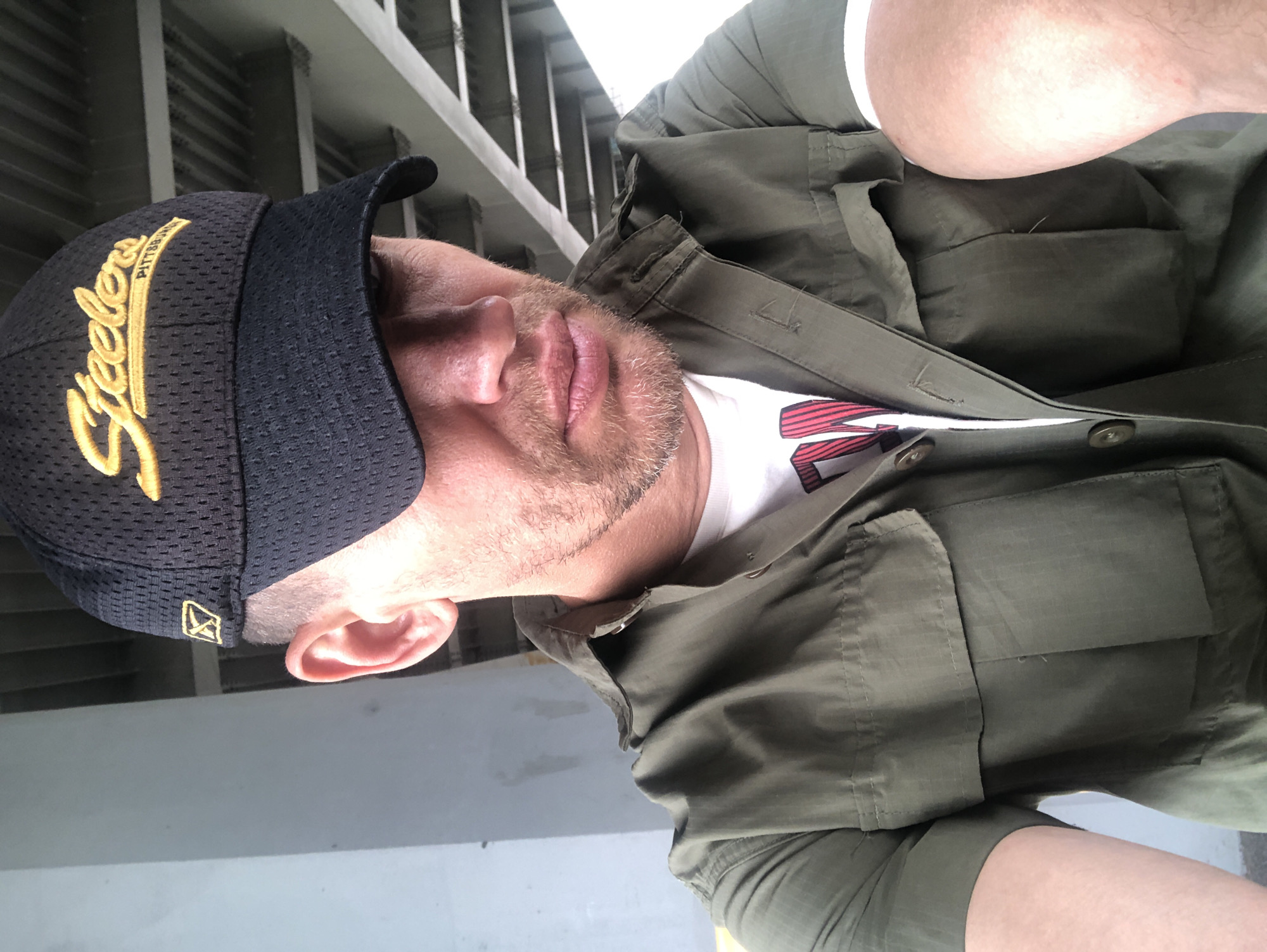 A man wearing a black Pittsburgh Steelers baseball cap and a green short-sleeved button-up shirt is pictured from the chest up, looking down and to the right. The man has a short beard and stubble. He is wearing a white t-shirt underneath his green shirt. The background appears to be an urban environment with concrete structures, possibly an underpass or bridge. The lighting suggests it is daytime. This image was taken in Saint Petersburg, Russia, likely near a public transportation hub or recreational area due to the concrete architecture.