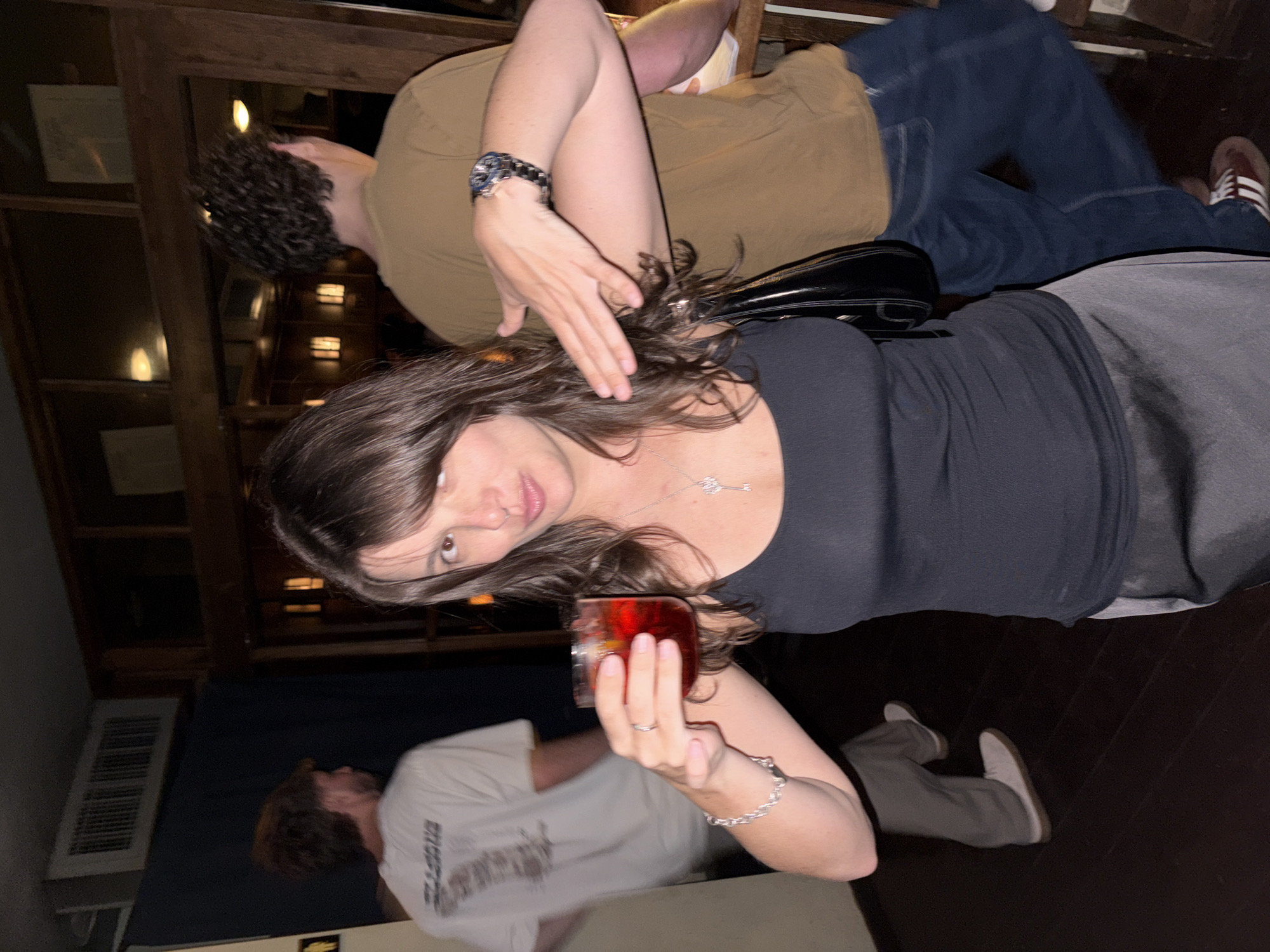 The image shows a woman holding a glass of red liquid. She wears a black top, gray pants, and a silver necklace and bracelet. A man with curly hair stands behind her, wearing a brown shirt, blue jeans, and a watch. Another man is visible in the background, wearing a white t-shirt and gray pants. The scene appears to be indoors, possibly in a bar or restaurant, with wooden paneling and a vent visible on the wall. The exact location in Tel-Aviv, Israel, cannot be determined from the image.