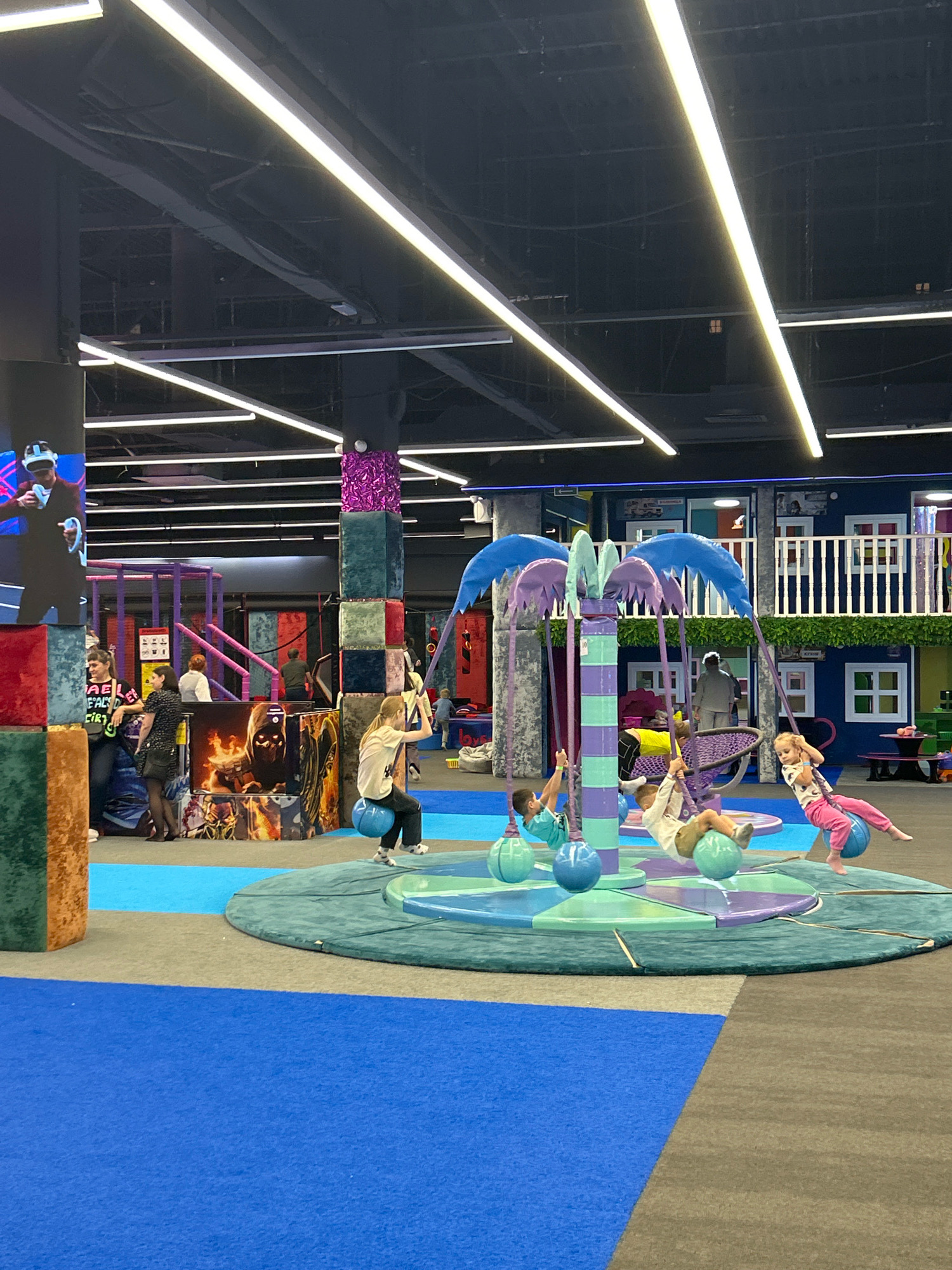 The image depicts an indoor children's play area located in Novokuznetsk, Russia. The space is characterized by bright, colorful flooring and various play structures. In the foreground, a circular platform features a unique swing set resembling a palm tree with purple and green elements. Four children are engaged with this swing set, each suspended from a spherical seat. One child, wearing pink pants and a white shirt, is actively swinging.
Behind the central play structure, a large, multi-colored pillar stands, adorned with blocks of varying textures and colors, including teal, purple, and metallic pink. To the left, a digital screen displays a person wearing a VR headset, suggesting interactive gaming or entertainment is available. Several adults are visible in the background, some observing the children and others interacting within the play area.
The environment is illuminated by long, linear fluorescent lights mounted on the dark ceiling. The background includes multi-level play structures with balconies and windows, painted in vibrant blues and whites, resembling a miniature town. The overall impression is a lively and engaging space designed for children's recreation.