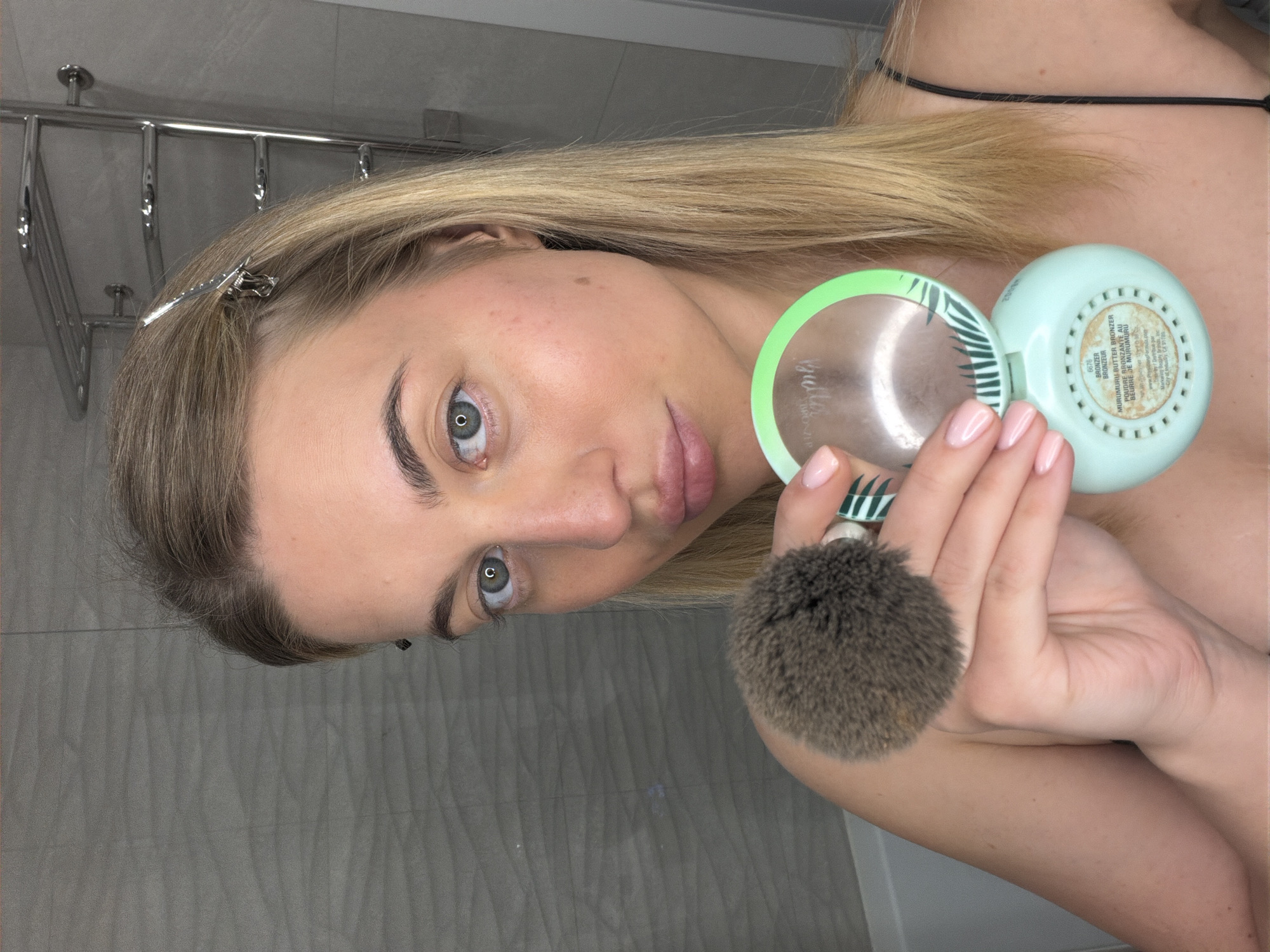 A close-up, angled portrait features a woman holding beauty products in an indoor setting, likely a bathroom or vanity area, in Sofiivska Borshchahivka, Ukraine. She has long, straight blonde hair, fair skin, and striking blue-green eyes. Her eyebrows are well-defined, and her lips have a soft pink tint. A silver hair clip secures her hair above her right ear.
She holds an open, light mint green compact and a fluffy, dark brown makeup brush in her left hand. The compact features a vibrant green rim with a palm leaf design, and its interior reveals a mirror-like surface and the product. The underside of the compact is also mint green with a grated texture. Visible text on the compact identifies the product as a "MURUMURU BUTTER BRONZER," with additional text in French "POUDRE BRONZANTE AU BEURRE DE MURUMURU," alongside "Bull," "2008円," "6676," "BRONZER," and "BRONZEUR." Her fingernails are neatly manicured with light pink polish.
The background consists of a grey tiled wall with a subtle textured pattern and a shiny, multi-tiered metal towel rack. The woman looks directly at the camera with a neutral expression, seemingly in the process of applying or demonstrating makeup.