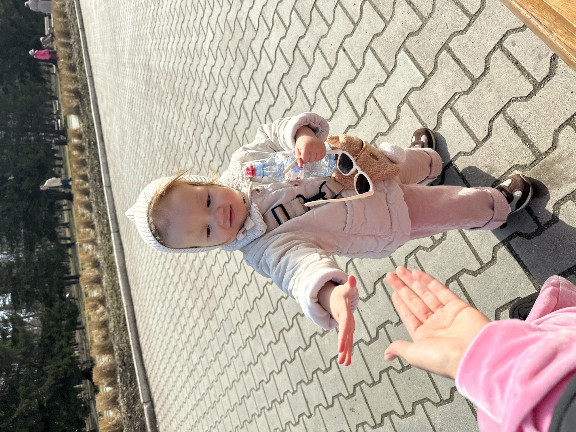 A baby wearing a white beanie, beige jacket, and pink pants, lying on a gray brick walkway. The baby holds a water bottle and a tan stuffed animal, and a pair of sunglasses is lying on the baby. A hand reaches towards the baby. Trees and benches are visible in the background. The location is Yekaterinburg, Russia.