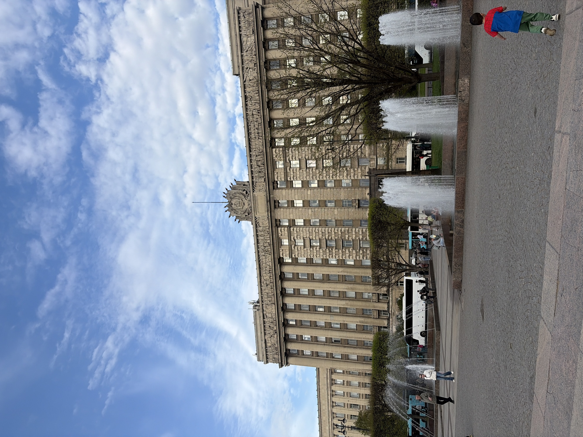 The photo shows a large building with a gray stone facade and many windows, partially obscured by bare tree branches. A long, multi-tiered fountain runs parallel to the building, with water jets shooting upwards. People are standing and walking around the fountain, and a boy wearing a blue and red shirt is running along the edge. A white bus is parked in the background, near the fountain. Above the building is a blue sky with scattered white clouds. The building and fountain are located in Moscow Square (Moskovskaya Ploshchad) in Saint Petersburg, Russia.