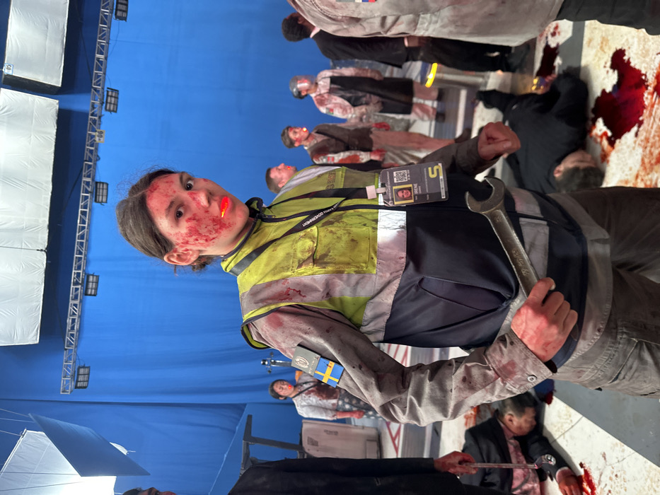 The image captures an indoor film set in Huangdao District, China. A prominent blue screen serves as the backdrop, with overhead metal truss structures and white reflective panels indicating studio lighting equipment. The floor is a light-colored surface extensively marked with numerous pools and spatters of simulated red blood.
In the foreground, a young individual with medium-length dark hair stands facing the viewer, positioned slightly to the left of the frame's center. The individual's face and neck are heavily covered in simulated blood, and a bright orange or red object is lodged in the mouth. Attire consists of a blood-stained, two-tone work jacket with a dark grey/navy lower section and sleeves, and a high-visibility yellow-green upper torso with reflective elements and pockets. A patch resembling the Swedish flag and a crest is affixed to the left sleeve. A black lanyard around the neck displays an ID badge with a photo of a woman named "IRENE" and a QR code. The individual holds a large, blood-stained silver-colored open-end wrench in the right hand.
Multiple additional individuals are visible in the midground and background, all bearing simulated bloodstains, suggesting their involvement in the same staged event. Some figures are standing, including one in a light-colored shirt, a male in a waistcoat, and another wearing an apron and face shield. On the floor, several figures lie motionless, surrounded by significant pools of simulated blood, one clearly identifiable as wearing a dark suit. The scene depicts the aftermath of a violent incident within a controlled production environment.
