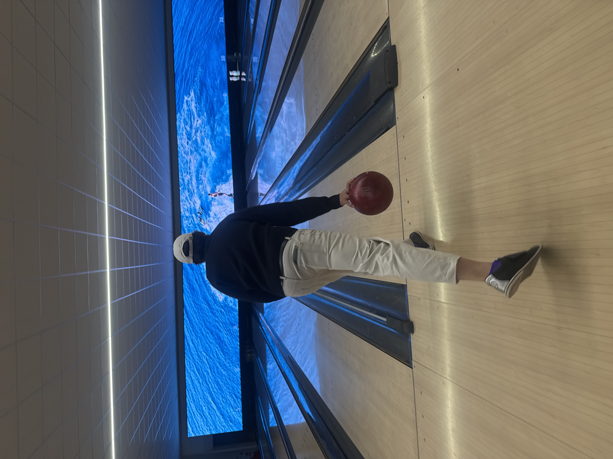 A person is captured mid-action in what appears to be a bowling alley. The individual is holding a red bowling ball, poised to release it down a wooden lane. They are wearing a black long-sleeved shirt, light-colored trousers, and bowling shoes. The bowling lane is surrounded by dark-colored gutters. In the background, a large screen displays a blue image, possibly depicting water or another dynamic scene. The adjacent wall is covered in square, grey panels illuminated by a bright light strip. This is very likely located in a bowling alley in Milan, Italy.