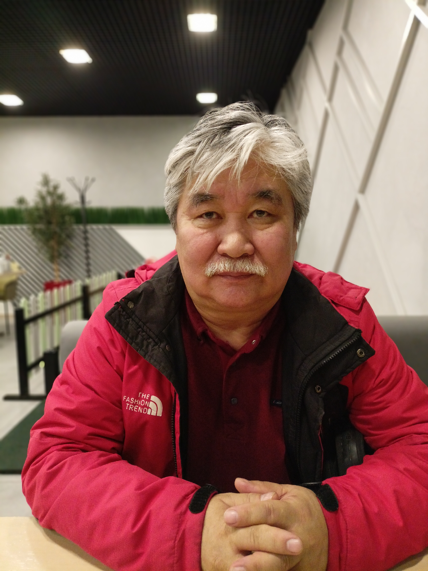 A middle-aged man is seated indoors at a table. He has graying hair and a mustache. He's wearing a red jacket over a red shirt. The jacket has a logo on the left side that says "THE FASHION TREND". His hands are clasped on the table. The background suggests an interior space with overhead lighting, and a partially visible decorative fence and greenery. Based on the setting, and available image information, this location is plausibly in Novosibirsk, Russia.