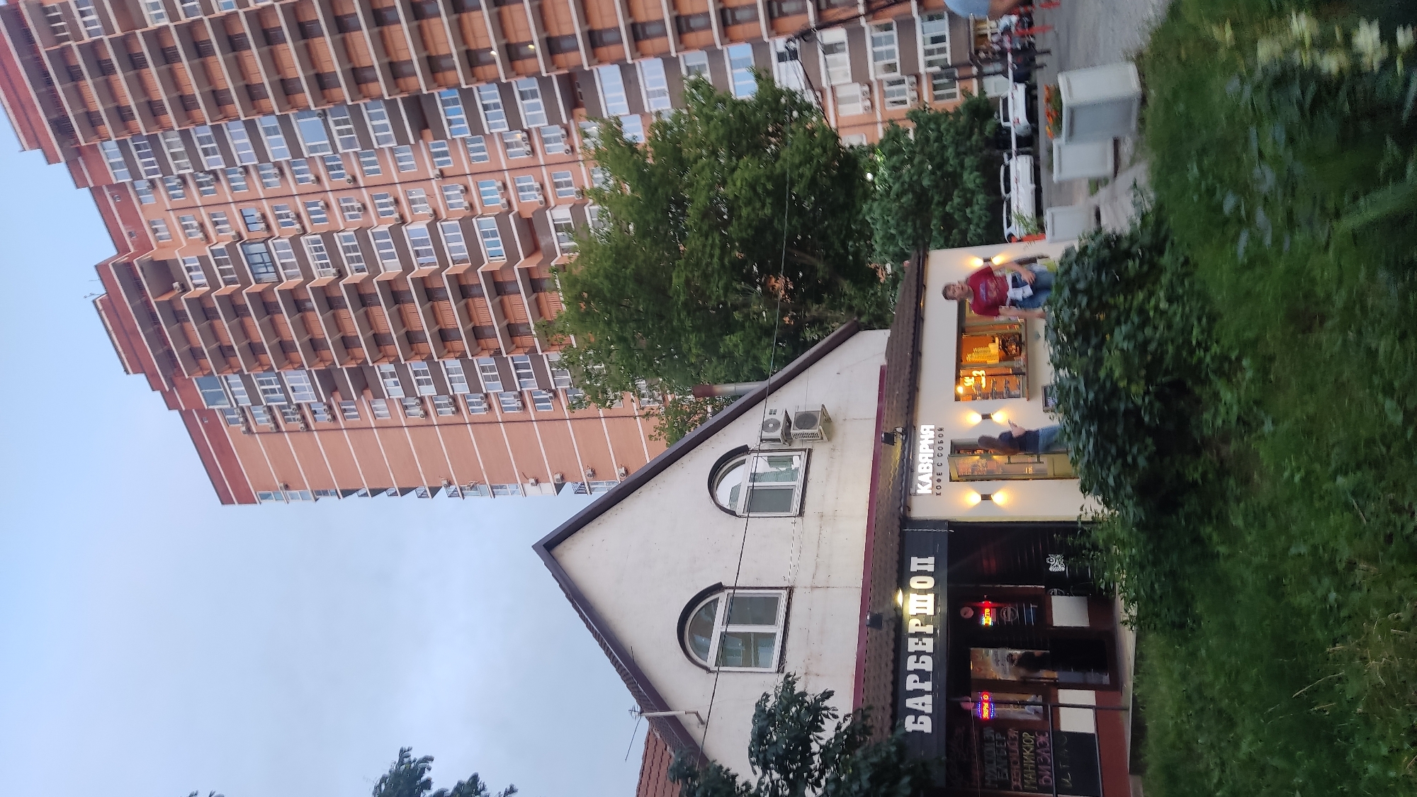 The image depicts a street scene in Krasnodar, Russia, featuring a low-rise building with two businesses and a tall, modern apartment building in the background. The low-rise building has a white facade and a dark-tiled roof. The business on the left has a sign that reads "БАРБЕРШОП" (Barbershop) in large, bold letters, with an open entrance showing a dimly lit interior. To the right of the barbershop is another establishment with a sign that says "КАВЯРНЯ" (Coffee Shop) and "КОФЕ С СОБОЙ" (Coffee To Go). This coffee shop has an open window counter where a person is visible.
In front of the establishments, there is a green, overgrown area with bushes and grass. A few people are visible; one person, a male in a red t-shirt, is standing near the coffee shop counter, and another person, a female in a blue shirt, is standing inside the coffee shop. In the background, the tall apartment building is a prominent feature, characterized by its brick facade and numerous balconies with glass railings. The sky is overcast, suggesting a cloudy day. Cars are parked on the street to the right of the coffee shop. The overall impression is an urban environment with a mix of older and newer architecture.