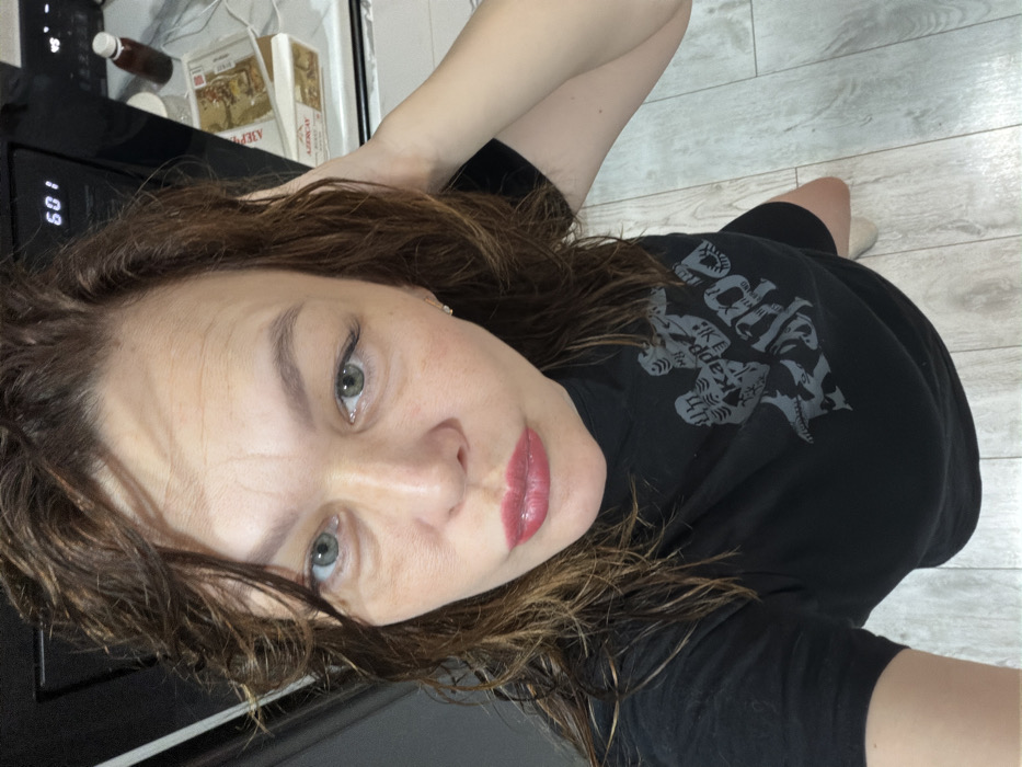A woman is depicted indoors, taking a selfie. She has medium-length wavy brown hair, light-colored eyes, and red-tinted lips. She is wearing a black t-shirt with a grey "Kappa" logo and other graphic elements. Her right hand is raised, partially obscured, appearing to touch the back of her hair. In the background to the upper right, a black microwave oven displays "60". Below the microwave, two boxes of "AZERÇAY" brand tea are visible on a countertop, alongside a small dark bottle with a light cap. The floor is covered with light-colored, plank-style flooring. The specific location within the city of Chaikovsky, Russia, cannot be determined from the image.