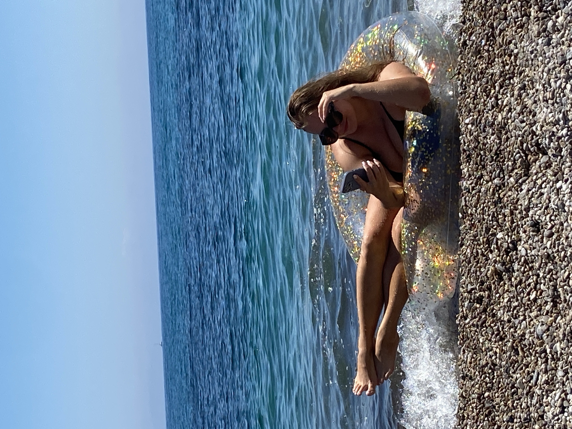 An outdoor, horizontally oriented photograph captures a woman relaxing by the sea on a bright day in Prijevor, Montenegro. The central subject is a woman, likely in her 20s or 30s, wearing a black bikini and sunglasses. She sits comfortably on a clear, inflatable ring filled with shimmering, iridescent glitter.
The woman is positioned at the water's edge, with the inflatable ring partially submerged in the calm, blue-green sea and partly resting on a pebbly beach. Her bare legs and feet extend into the shallow water, where small waves gently lap. She is holding a dark-colored smartphone in both hands, looking down at its screen, suggesting she is engaging with it. Her hair is down, and she appears relaxed.
The background features the expansive, tranquil sea stretching to a clear horizon under a light blue sky. A small, distant boat can be seen on the horizon line. The foreground consists of distinct, light-brown pebbles, indicating a pebbly rather than sandy beach. The strong, natural light suggests it is daytime, likely mid-morning or afternoon. No visible text is present in the image.