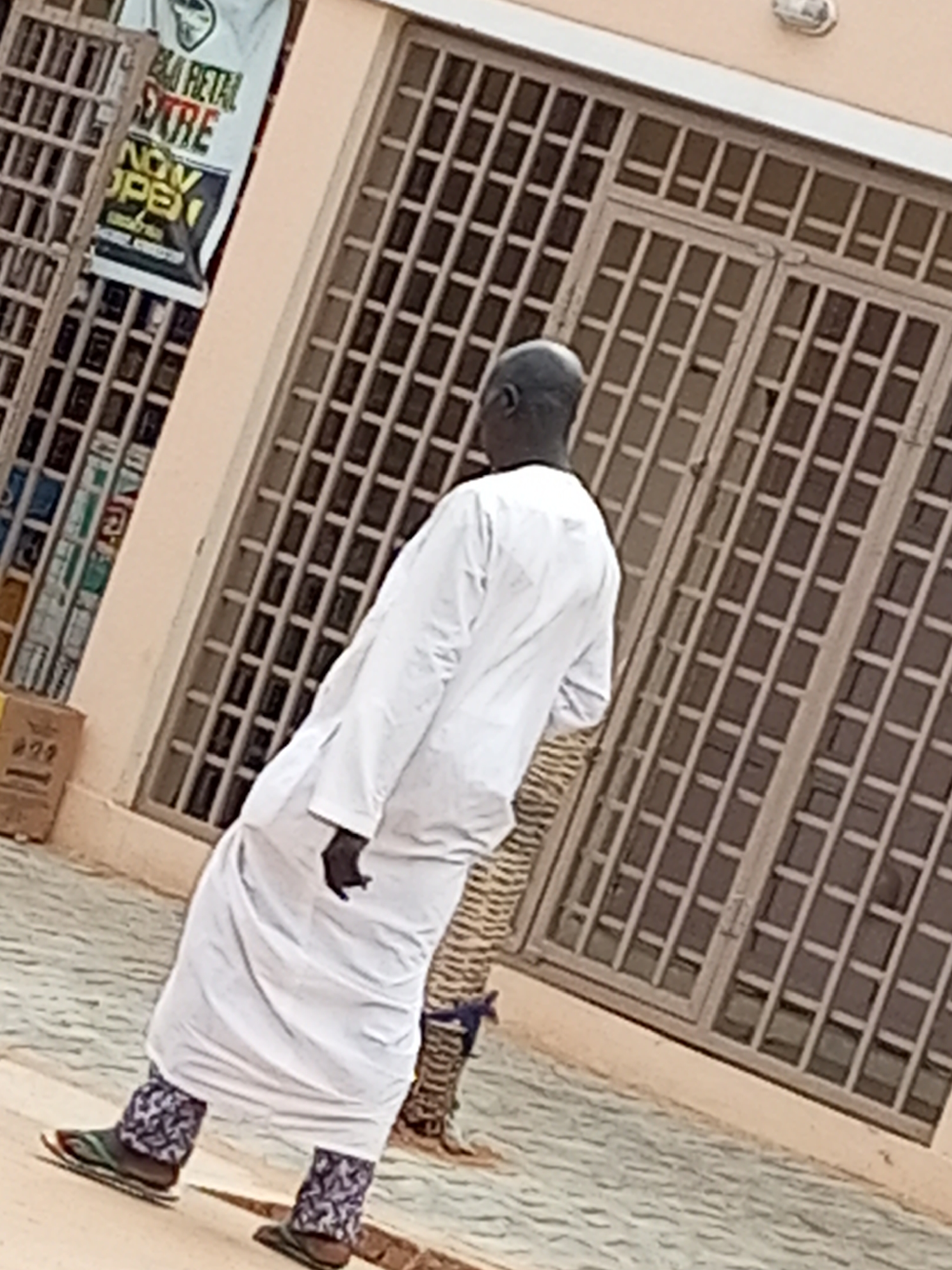 A man in a white robe walks on a paved street in front of a building with metal-barred windows. The building appears to be a shop, as evidenced by a banner on the left side of the image that reads "RETAIL EXTRA NOW OPEN". The man is facing away from the camera and appears to be walking to the left. The street is made of grey interlocking pavers, and there is a small potted plant to the right of the man. The sky is not visible in the image.