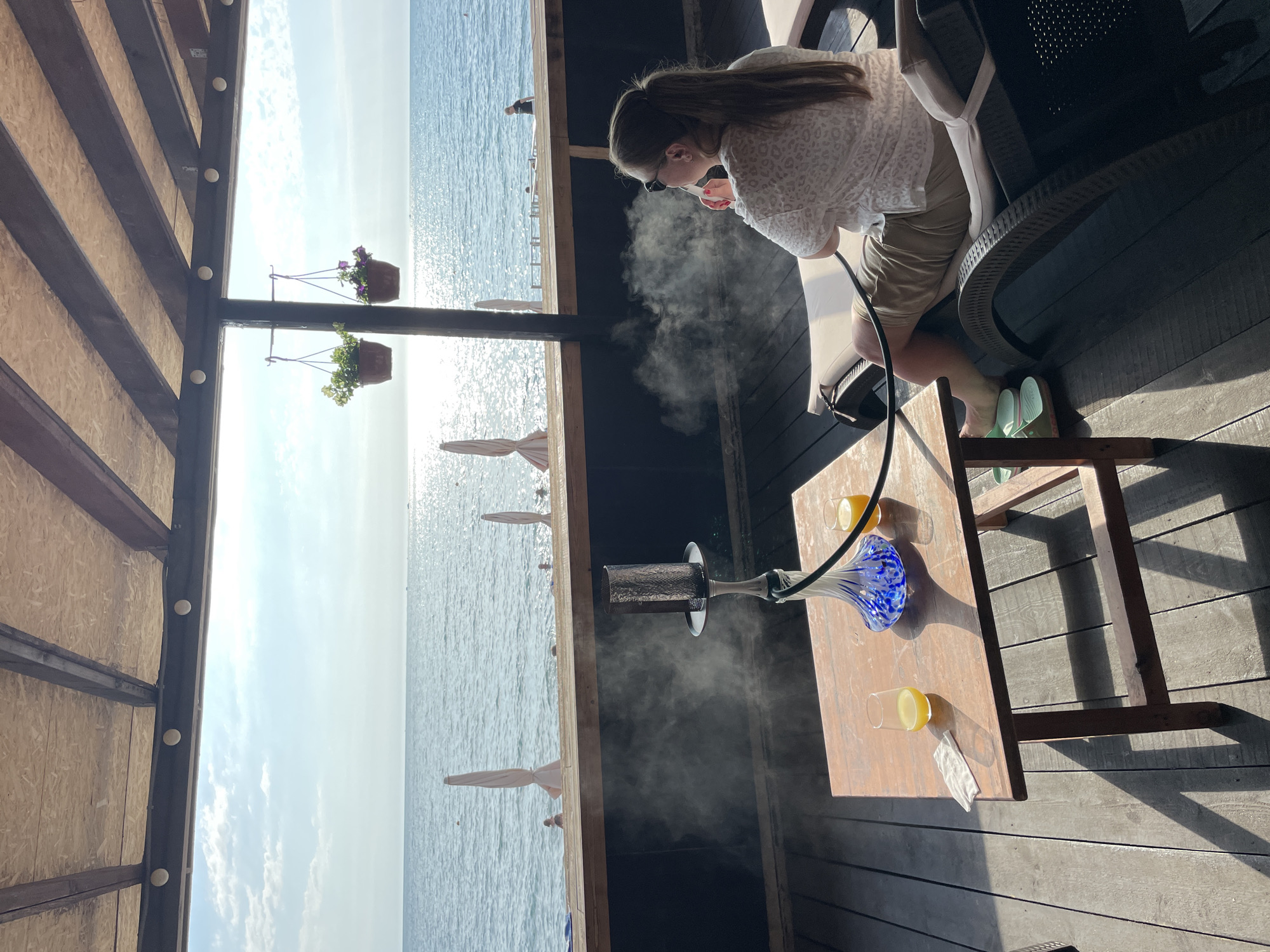 In Sochi, Russia, a woman is relaxing on a lounge chair on a wooden deck, overlooking the Black Sea. She is wearing a white leopard-print shirt and khaki shorts, with sunglasses perched on her head. She is holding a hookah hose, exhaling a cloud of smoke. In front of her, on a wooden table, are two glasses of orange juice and a small white napkin. A hookah pipe sits on the table, its blue and white base contrasting with the dark wood. In the background, the sea stretches out to the horizon, with a few beach umbrellas and people visible along the shore. The sky is a clear blue with scattered clouds, and the sun reflects brightly off the water. The scene is dappled with sunlight and shadow, creating a relaxed atmosphere.