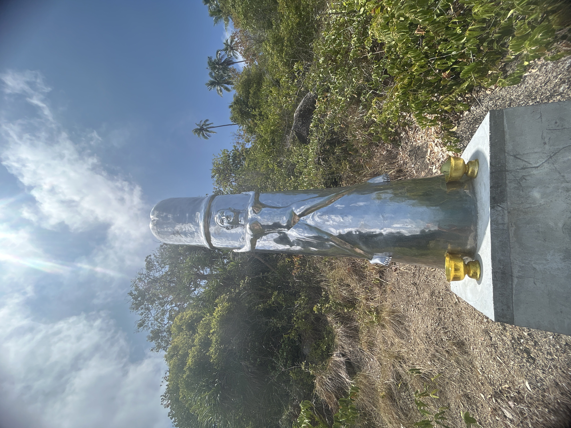 A vertically oriented image captures a tall, slender, highly reflective metallic statue positioned outdoors. The statue, possibly made of polished stainless steel, depicts a humanoid figure with visible facial features and what appears to be robes or draped clothing. Its surface reflects the bright blue sky and surrounding vegetation. The statue is mounted on a grey concrete pedestal, and golden, goblet-like elements are visible at its base where it meets the pedestal, suggesting decorative or structural support.
The setting is a tropical environment. Behind the statue, a hillside rises, covered with a mix of dense green foliage, shrubs, and patches of dry, brown grass. Several distinct palm trees are visible against the sky in the upper left and middle-right background. The sky is clear blue with scattered white clouds, and bright sunlight causes a noticeable lens flare in the upper left quadrant of the image. The overall impression is a sunny day. No human activity is depicted, nor is any visible text present. The scene captures a tranquil moment with the striking metallic figure contrasting against the natural tropical landscape. Given the location context, this statue is likely a point of interest or religious monument in Baan Thong Nang, Thailand.