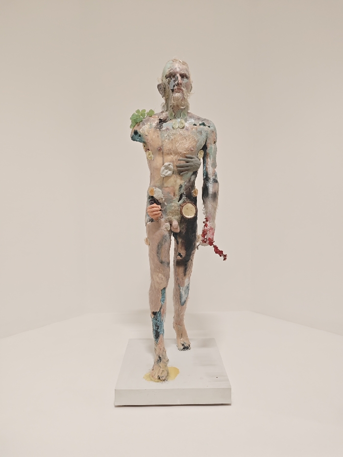 An upright, full-body sculpture of a male figure stands centrally within a brightly lit, minimalist gallery space, against plain off-white walls that form an interior corner. The figure is positioned on a white rectangular plinth, with a yellow, viscous-looking substance pooling around its left foot. The sculpture presents a textured, multi-colored surface, incorporating hues of pale pink, beige, blue, black, white, and red, suggesting diverse materials or a state of decay. The head features a bearded male face. The left arm is absent from the shoulder, where a cluster of green, clover-like forms is attached. The torso displays several embedded elements: a human-like hand presses against the chest on the viewer's left, and a smaller, lighter-toned hand emerges near the groin on the same side. Multiple circular and irregularly shaped objects, some resembling buttons or shell fragments, are visible across the chest and abdomen, including one that suggests a clock face or compass. The right arm extends downward, from which a cluster of red, organic-looking material hangs. The figure is depicted in a walking pose, with the left foot forward. The specific location within the city of Paris, France, cannot be determined from the visual information provided.