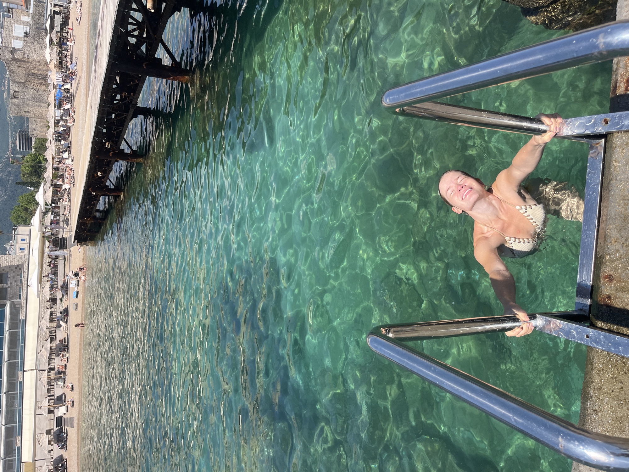 A woman in a light-colored bikini is climbing out of the crystal-clear, turquoise water using a metal ladder. She is smiling and squinting, possibly from the bright sun. The water is transparent, allowing a view of the rocky bottom and sunlight patterns on the surface. To the left of the water is a sandy beach lined with lounge chairs and umbrellas, with buildings and a stone wall in the background. A pier extends into the water from the left side of the image, supported by dark metal beams. The scene suggests a sunny day at a beach resort in Budva, Montenegro. The overall mood is relaxed and joyful.