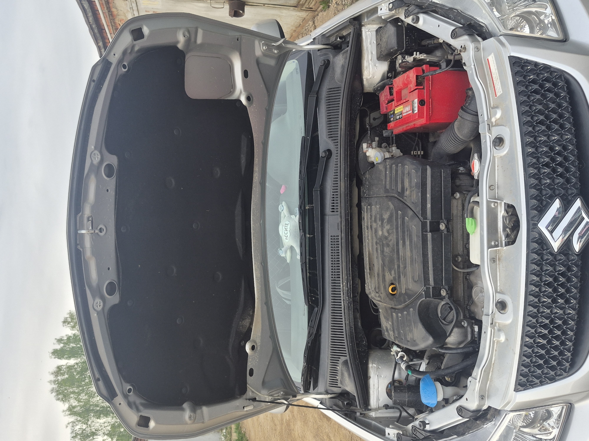 The image shows the open hood of a silver Suzuki Swift. The engine compartment is visible, with the engine, battery (with a red casing), and various hoses and components. The windshield wipers are in the parked position. A small plush toy is visible on the dashboard behind the windshield. The environment appears to be outdoors, possibly near a building, with some vegetation visible in the background. The location is Unknown, Unknown.