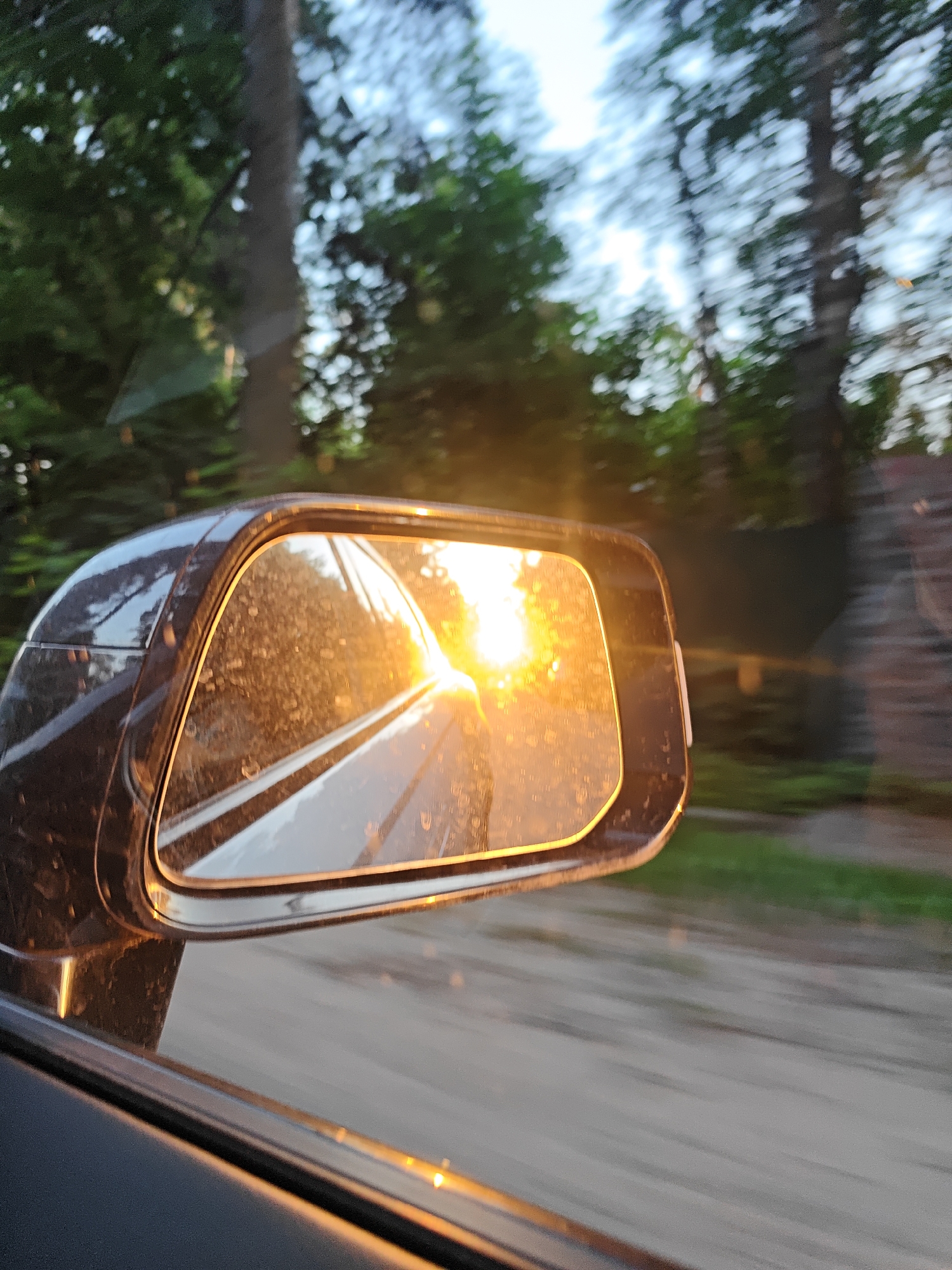 This image captures a close-up perspective of a car's passenger-side exterior mirror and its immediate surroundings in Kratovo, Russia. The sun, likely either rising or setting, is directly reflected within the mirror, creating an intense, golden-orange light flare that dominates the reflective surface and casts lens flares across the image. The mirror itself is visibly smudged and covered with small particles of dust or dirt. A dark, possibly metallic, portion of the car's bodywork forms the frame around the mirror and extends into the lower left corner of the image, where a segment of the vehicle's interior door panel is also visible. Through the car window and beyond the mirror, the background consists of out-of-focus green foliage from numerous trees, exhibiting motion blur consistent with a moving vehicle. Patches of light sky are discernible between the tree canopy. Below the tree line, a blurred grey-brown expanse suggests a road or dirt track, with hints of green roadside vegetation. On the far right, a dark, indistinct structure, possibly a building or fence, is partially visible and also affected by motion blur. The overall lighting is warm, with strong backlighting from the sun. No human figures are present in the scene.