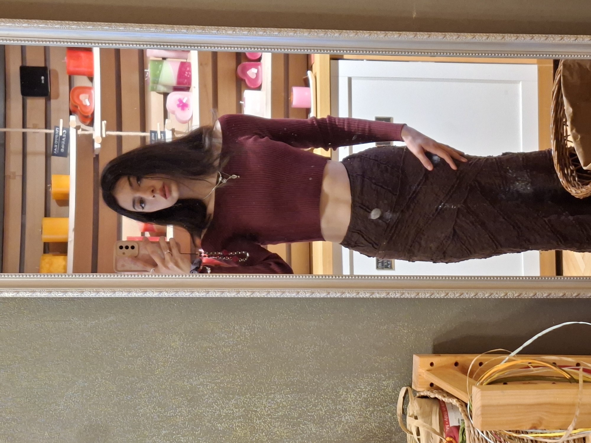 The image is a full-body mirror selfie of a young woman, captured with the image rotated 90 degrees clockwise. In the reflection, she stands upright, looking into the smartphone she holds in her right hand. She has long, dark hair, dark eyes, and a neutral expression. She is wearing a fitted, ribbed maroon crop top with long sleeves, revealing her midriff, and a dark brown, textured, possibly suede or corduroy, long skirt. A delicate silver-colored necklace with small charms adorns her neck, and a ring is visible on her right hand.
The setting appears to be an interior space, likely a gift or craft shop in Sofia, Bulgaria. Behind the woman, reflected in the mirror, are wooden shelves displaying various colorful candles. These include red, yellow, and orange rectangular block candles, as well as heart-shaped candles in pink and red, and a multi-colored green and pink swirled candle. Small wooden signs are visible on the shelves, one clearly reflecting "CANDLE" and another partially visible, reflecting "CERTIFIED". A white door is also reflected on the right side of the mirror. In the upper right corner of the mirror's reflection, a light-colored woven basket is partially visible.
In the actual room, framing the mirror in the foreground, a muted greenish-brown textured wall is visible. Below the mirror, a wooden display unit holds craft supplies, including spools of colorful ribbons or threads in shades of yellow, white, red, and green, along with another woven basket. The mirror itself has an ornate, silver-colored frame. The lighting is bright and even, suggesting daytime.