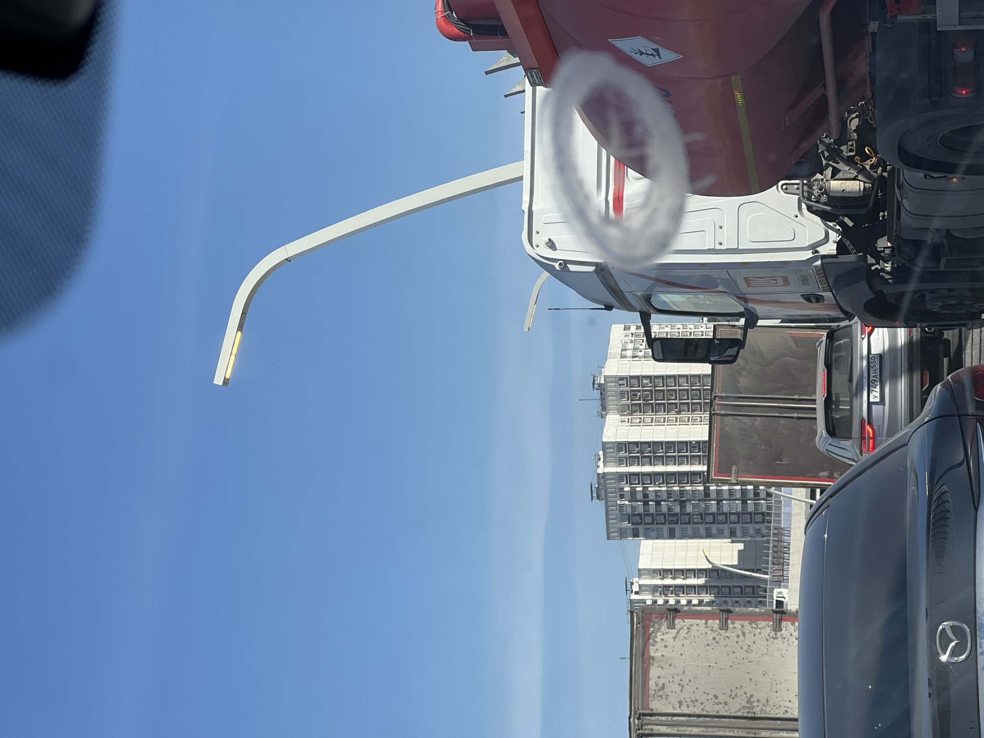 The image shows a traffic scene in Moscow, Russia, with a clear blue sky in the background. Several vehicles are visible, including a red and white tanker truck, a gray car with license plate Y749AH55, and a black Mazda. In the distance, there are tall residential buildings. There is a white, curved street light visible.