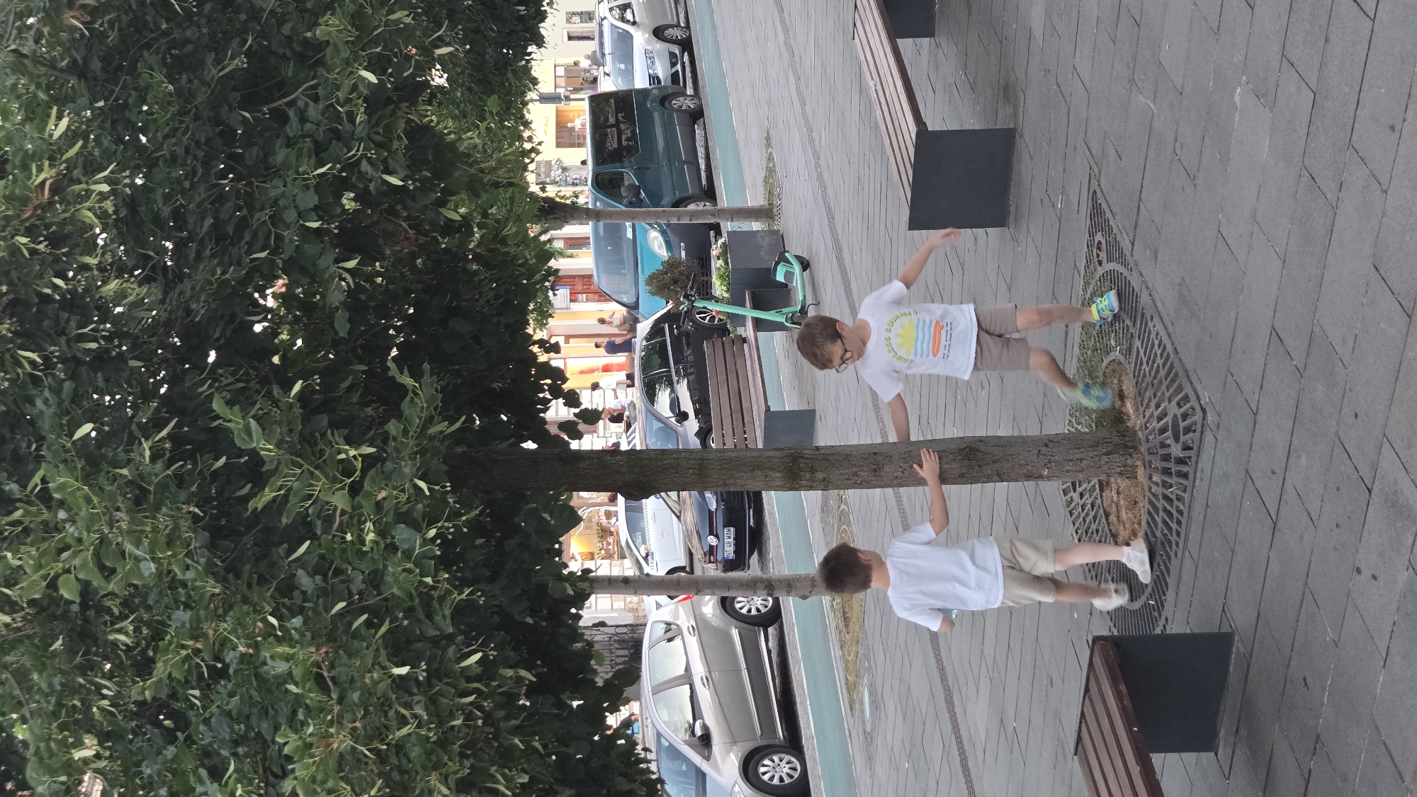 Two young boys, both wearing white t-shirts and shorts, are standing by a tree on a paved walkway. The boy on the left is wearing tan shorts and white shoes, while the boy on the right is wearing khaki shorts, blue and green shoes, and glasses. He is also wearing a t-shirt with a graphic that says "COLLEGE SUMMER." The boys have their hands on the tree. There are several cars and a teal van parked on the street in the background. A teal scooter is parked on the walkway to the right of the tree. Benches are present on the walkway. The scene appears to be in a European city, likely Cluj-Napoca, Romania, given the context of the original request.