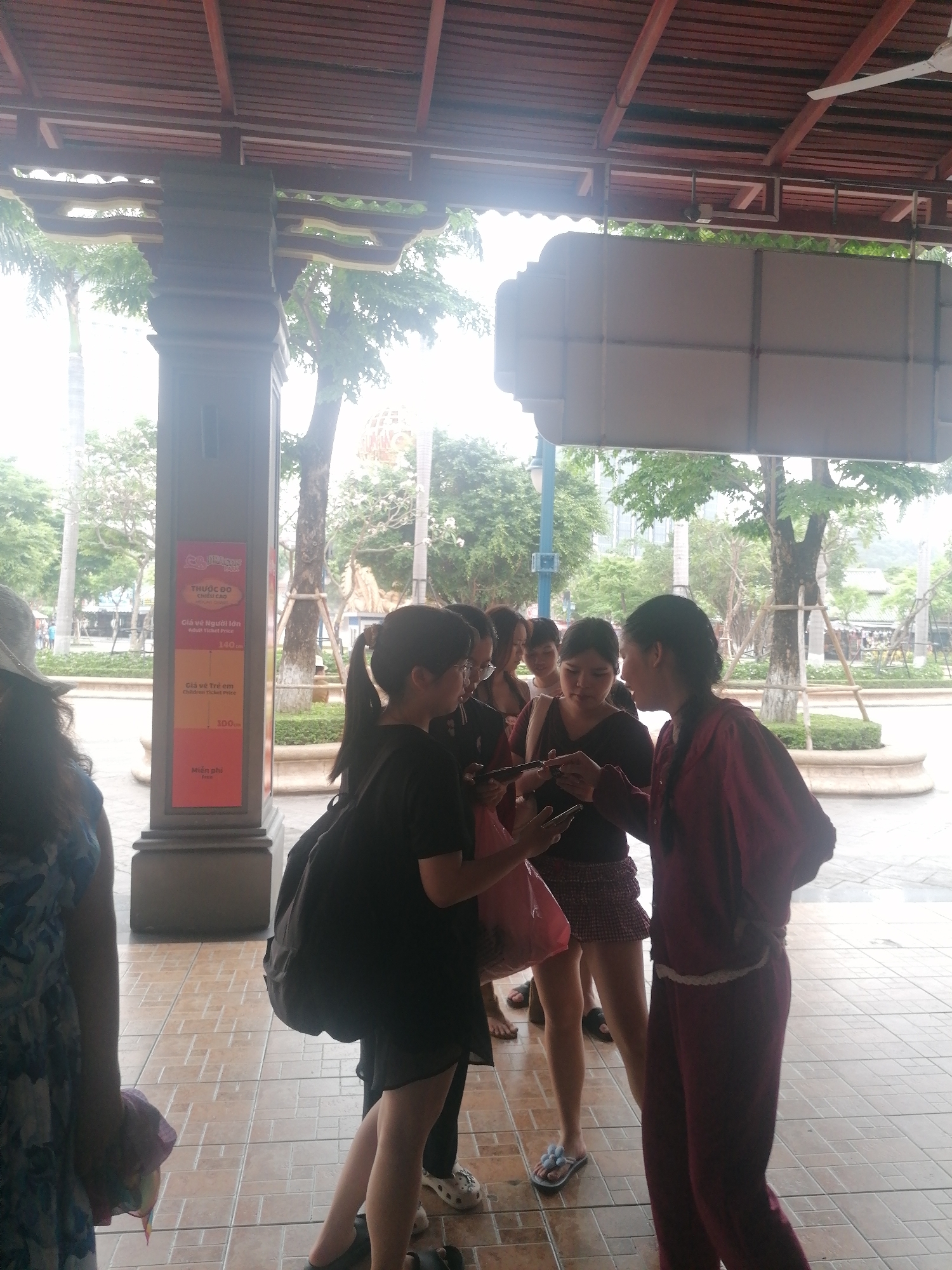 A group of approximately six to seven young women is gathered under a covered outdoor area, possibly an entrance or waiting point for an attraction. They are casually dressed, with some looking at their mobile phones. One woman in a dark red tracksuit points towards a phone screen held by another woman in a black shirt, suggesting they are discussing or sharing information from the device. The women are positioned on a tiled floor beneath a wooden slatted roof structure, with an ornate pillar nearby.
On this pillar, a brightly colored sign is displayed, primarily in red, orange, and yellow segments. The text, in Vietnamese and English, indicates a "THƯỚC ĐO CHIỀU CAO" (Height measurement). Below this, "Giá vé Người lớn" (Adult Ticket Price) is listed as "140", and "Giá vé Trẻ em" (Children Ticket Price) as "100". The bottom segment reads "Miễn phí" (Free). These details suggest the location is likely an amusement park, theme park, or another paid attraction where height and age determine ticket prices.
In the background, lush green trees and other foliage are visible under bright daylight, hinting at an outdoor recreational environment. A large, blank signboard is suspended from the roof structure. The scene is set in Bai Chay Ward, Vietnam.